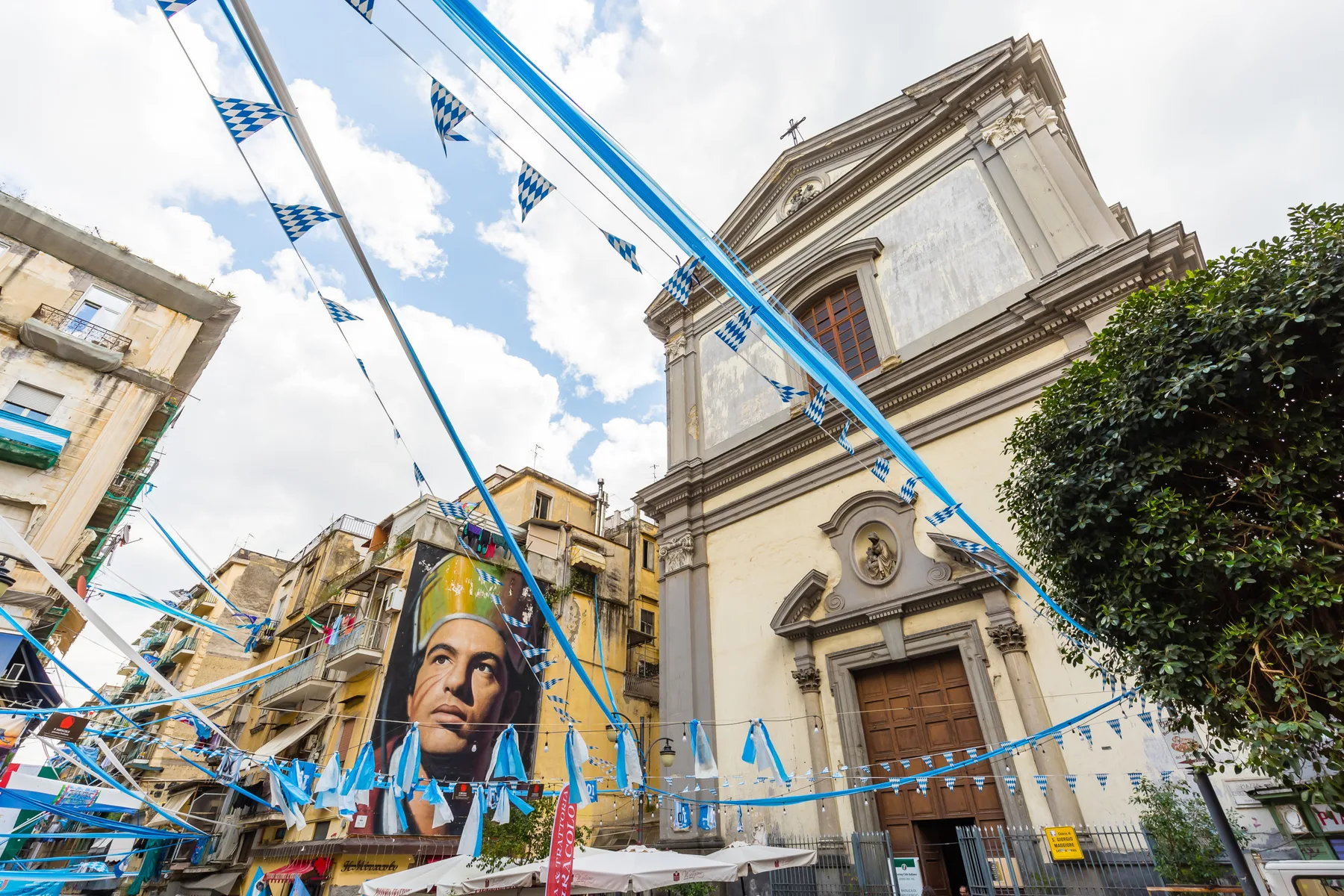 La fachada de una iglesia está junto a un edificio con un mural, con banderines azules sobre la calle.