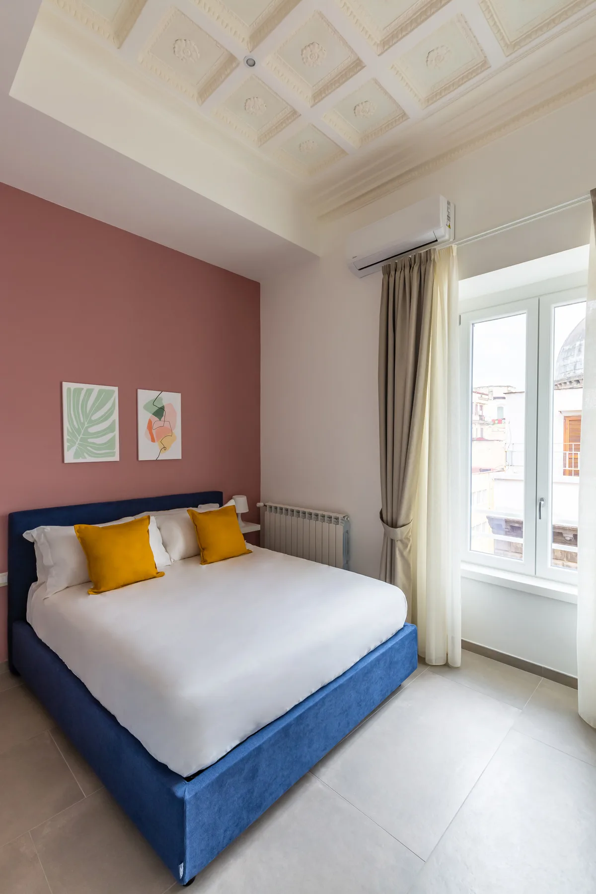 Un dormitorio con una cama azul, ropa de cama blanca y una ventana con cortinas.