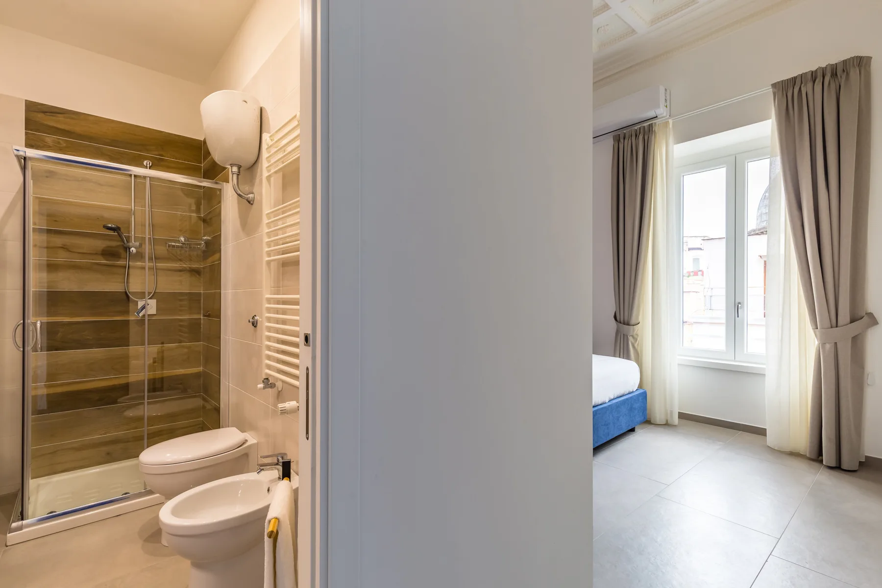 Un baño con ducha de vidrio junto a una habitación luminosa con una ventana con cortinas.
