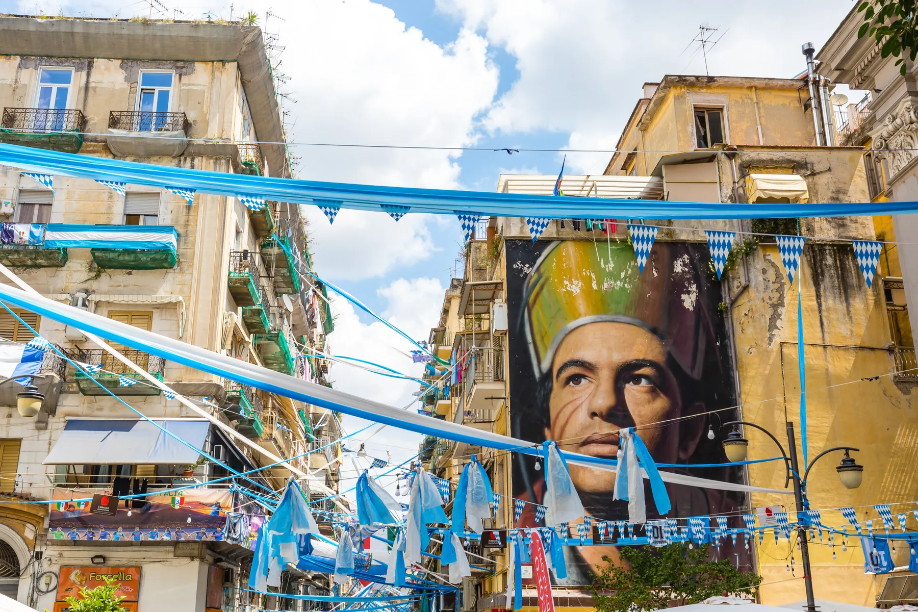 Un gran mural cubre la fachada de un edificio bajo adornos azules tendidos sobre la calle.