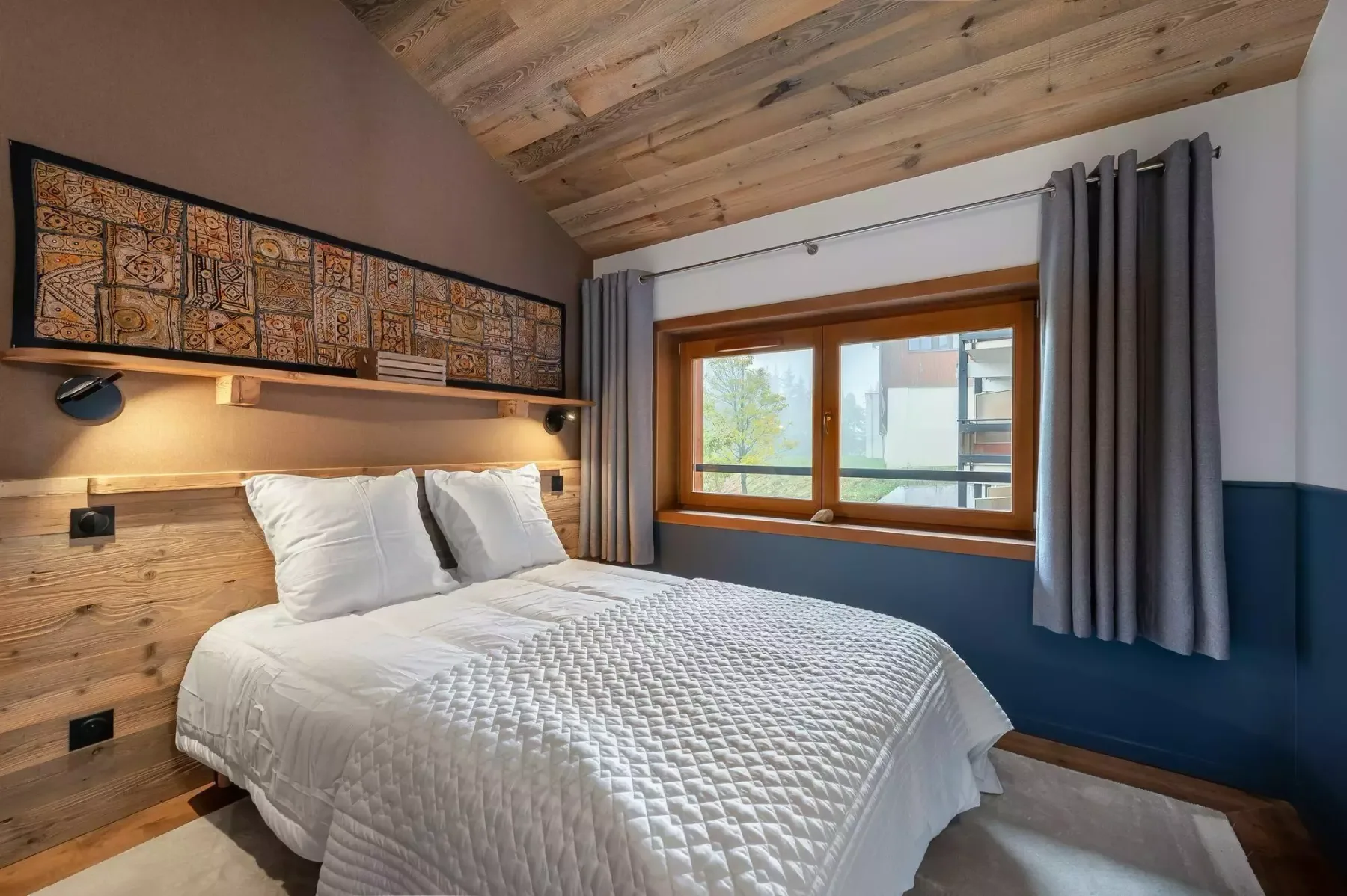 Dormitorio con una cama bajo un techo de madera y una ventana amplia.