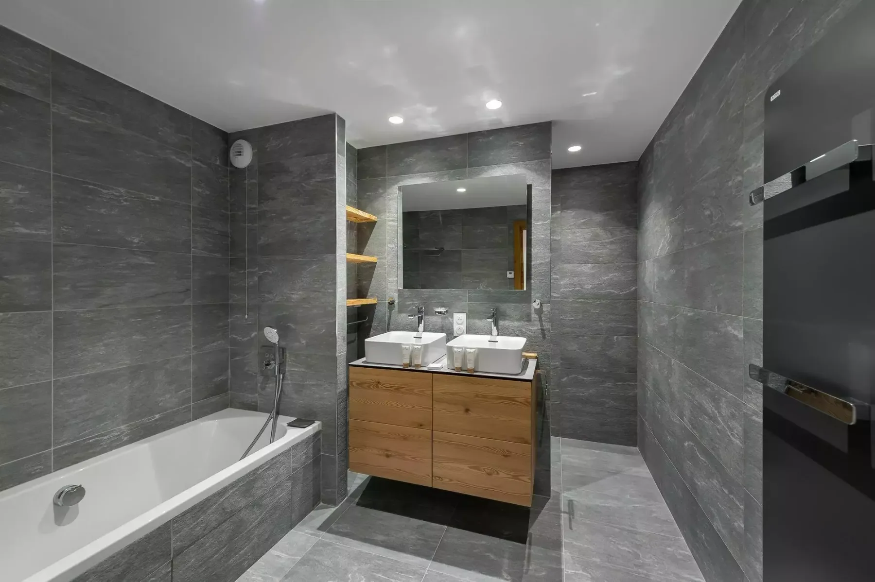 Baño moderno con bañera, lavabo doble y azulejos grises.
