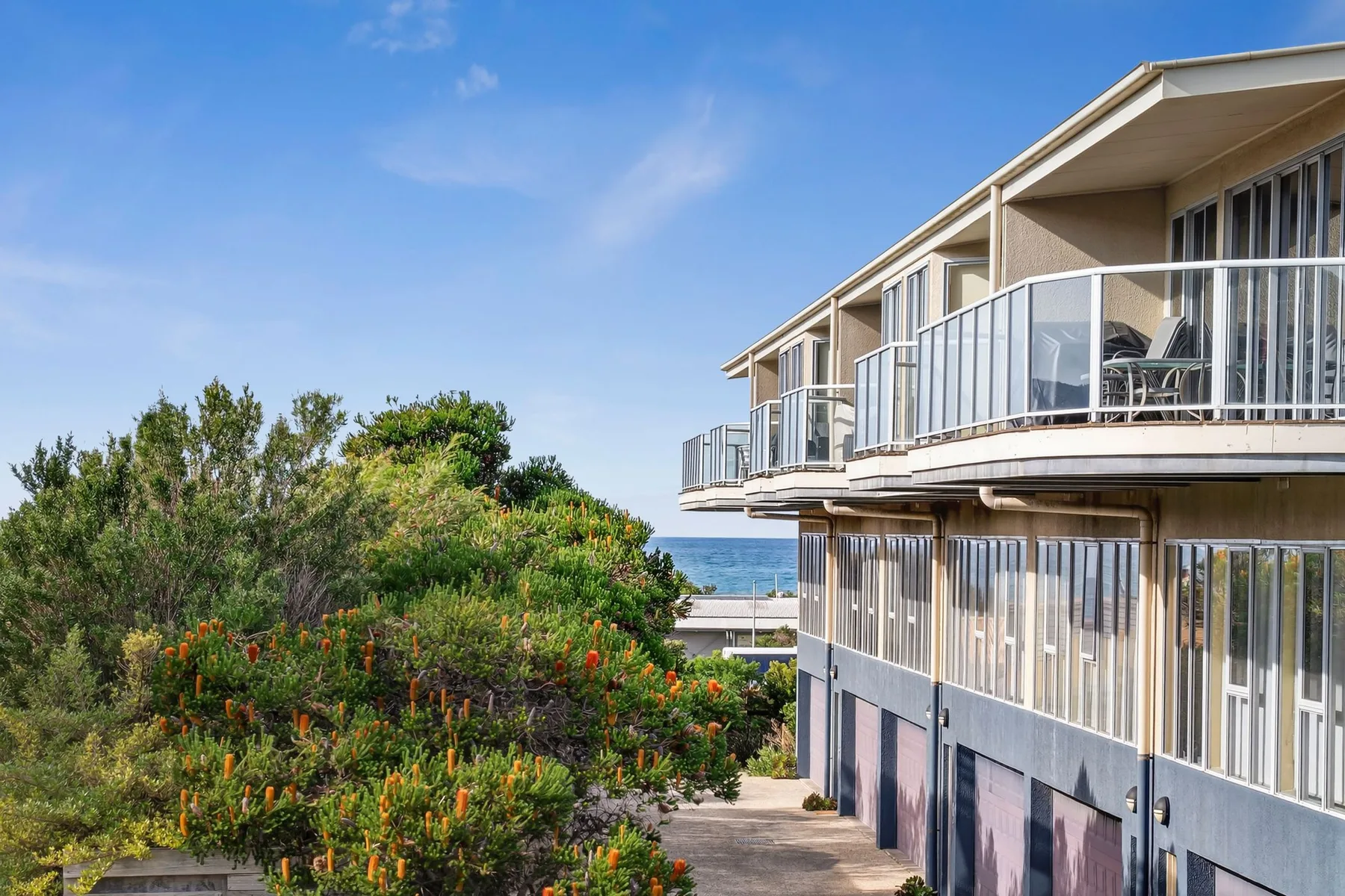A modern multi-story building with glass balconies stands beside landscaped greenery.