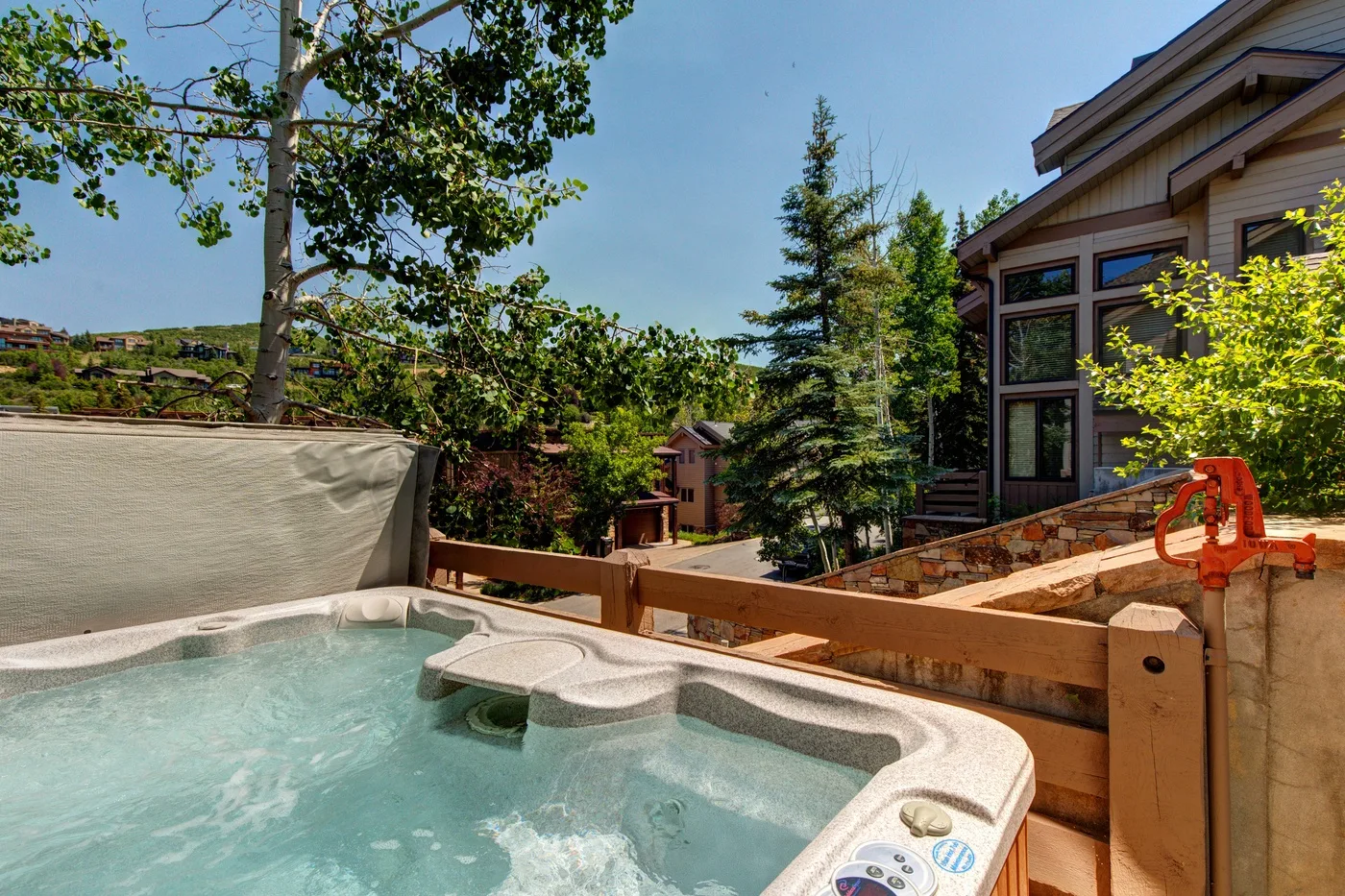 Un jacuzzi exterior está junto a una terraza de madera, con una casa y árboles cerca.
