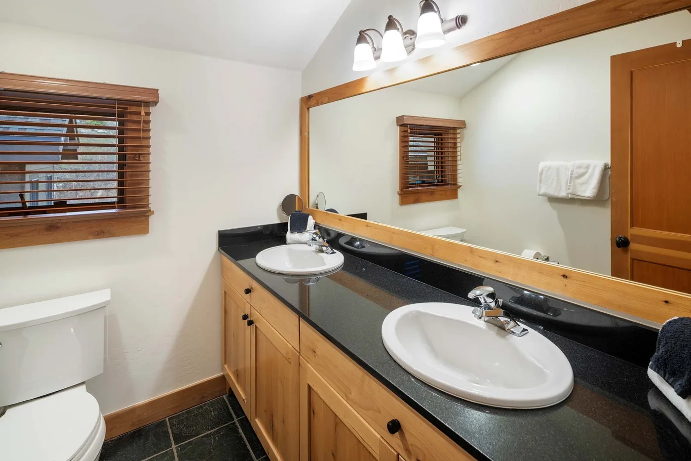 Tocador de baño con un espejo amplio, gabinetes de madera y una ventana con persianas.
