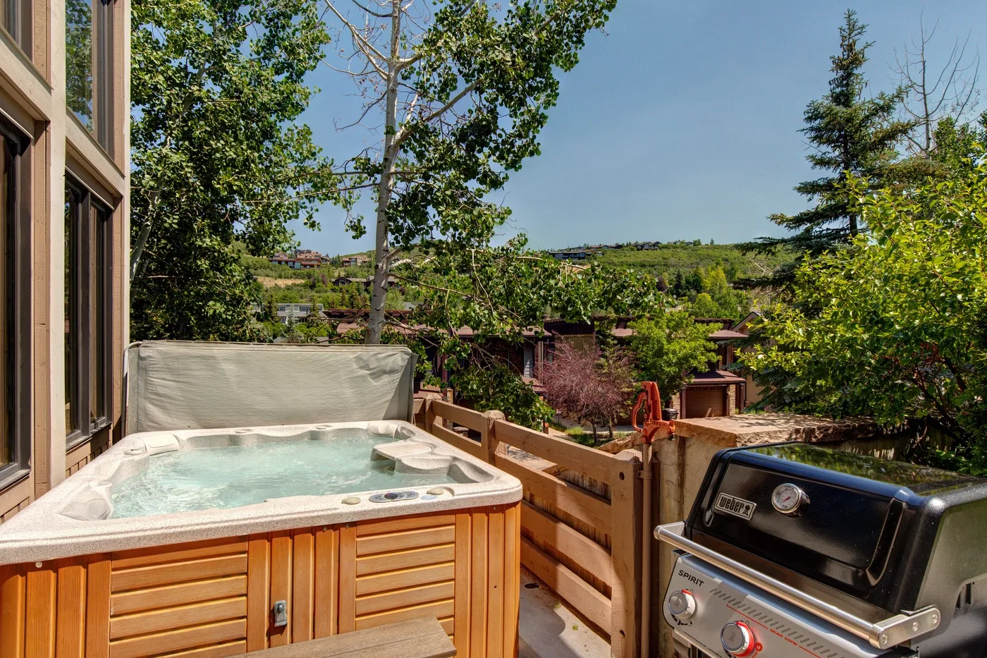 A hot tub sits on a wooden deck beside a barbecue grill, with trees beyond.