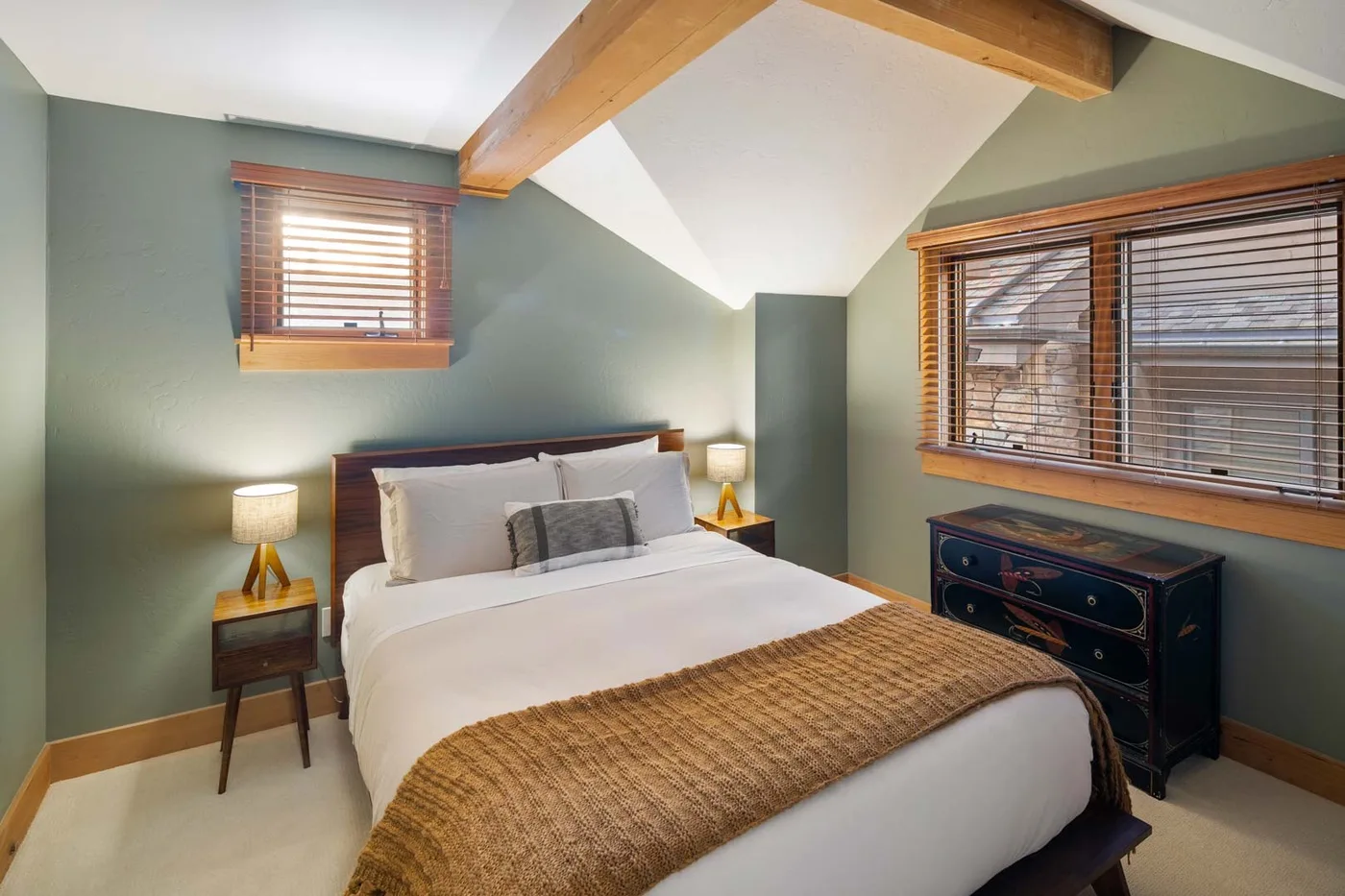 Un dormitorio con una cama, mesitas de noche y ventanas con persianas de madera.