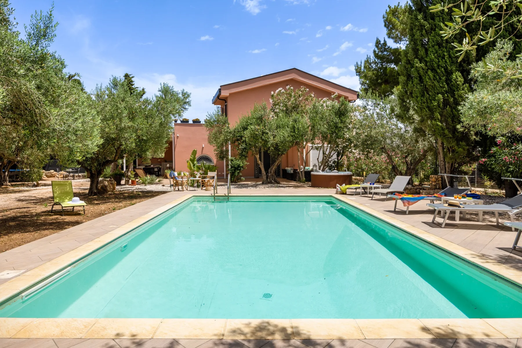 An outdoor swimming pool sits in front of a house, with trees and loungers nearby.