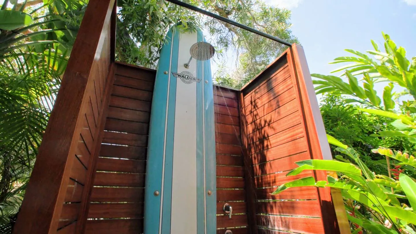 Una ducha exterior está entre paneles de madera, rodeada de vegetación tropical.