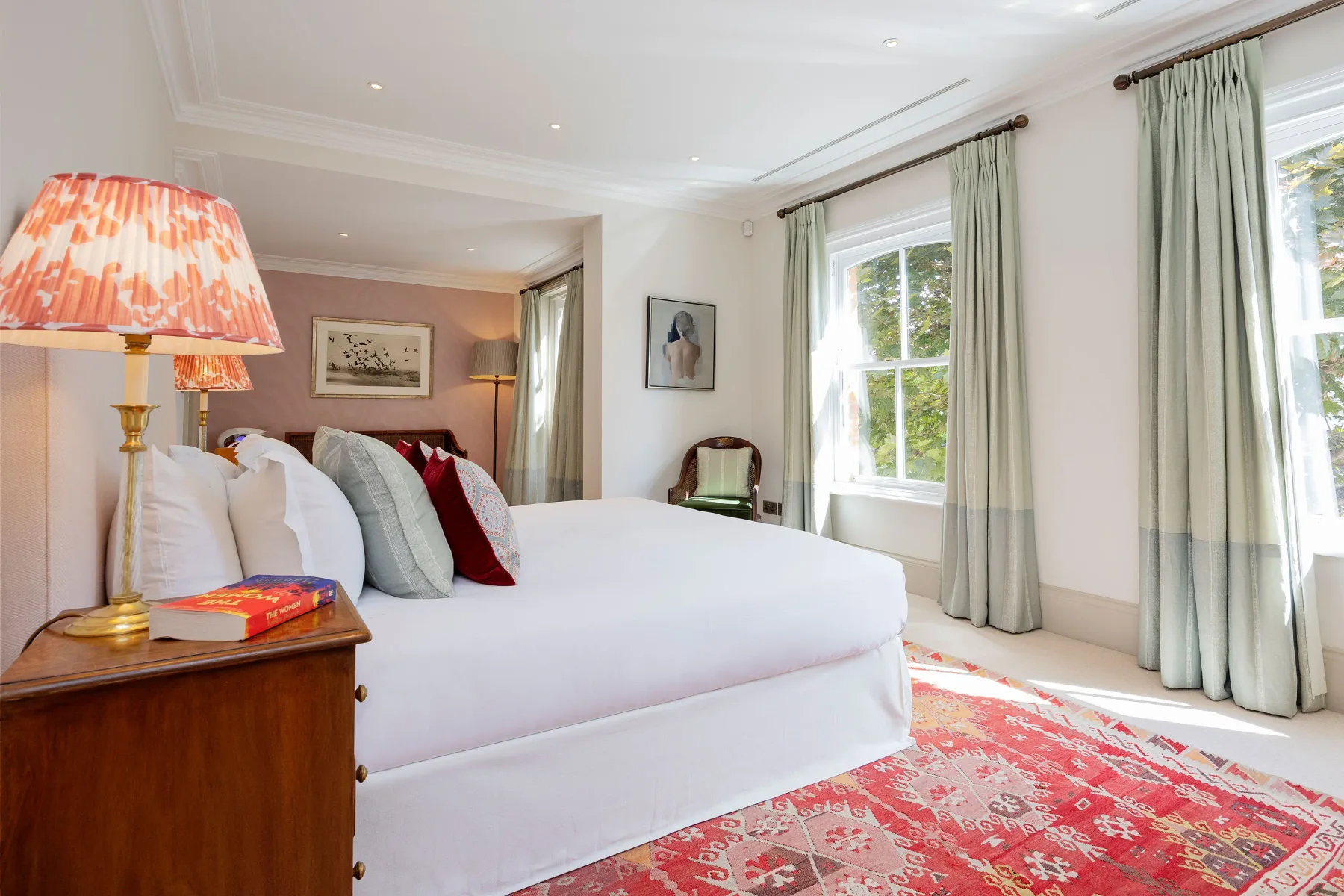 Un dormitorio luminoso con una cama grande, una lámpara de noche y ventanas altas con cortinas.