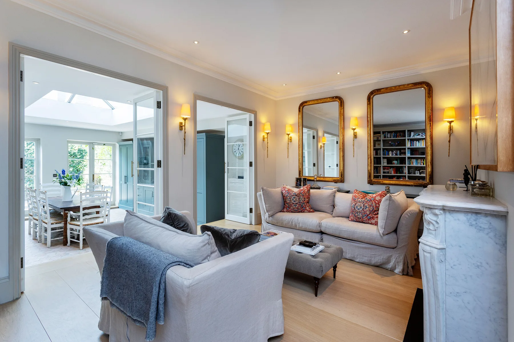 A bright living room with seating, a bookcase, and a dining area visible through the doorway.