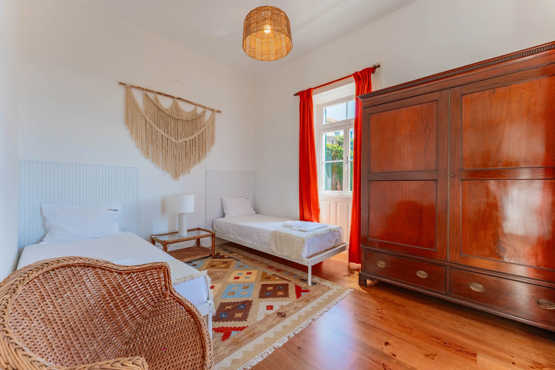 Un dormitorio luminoso con dos camas, un armario de madera y cortinas rojas.
