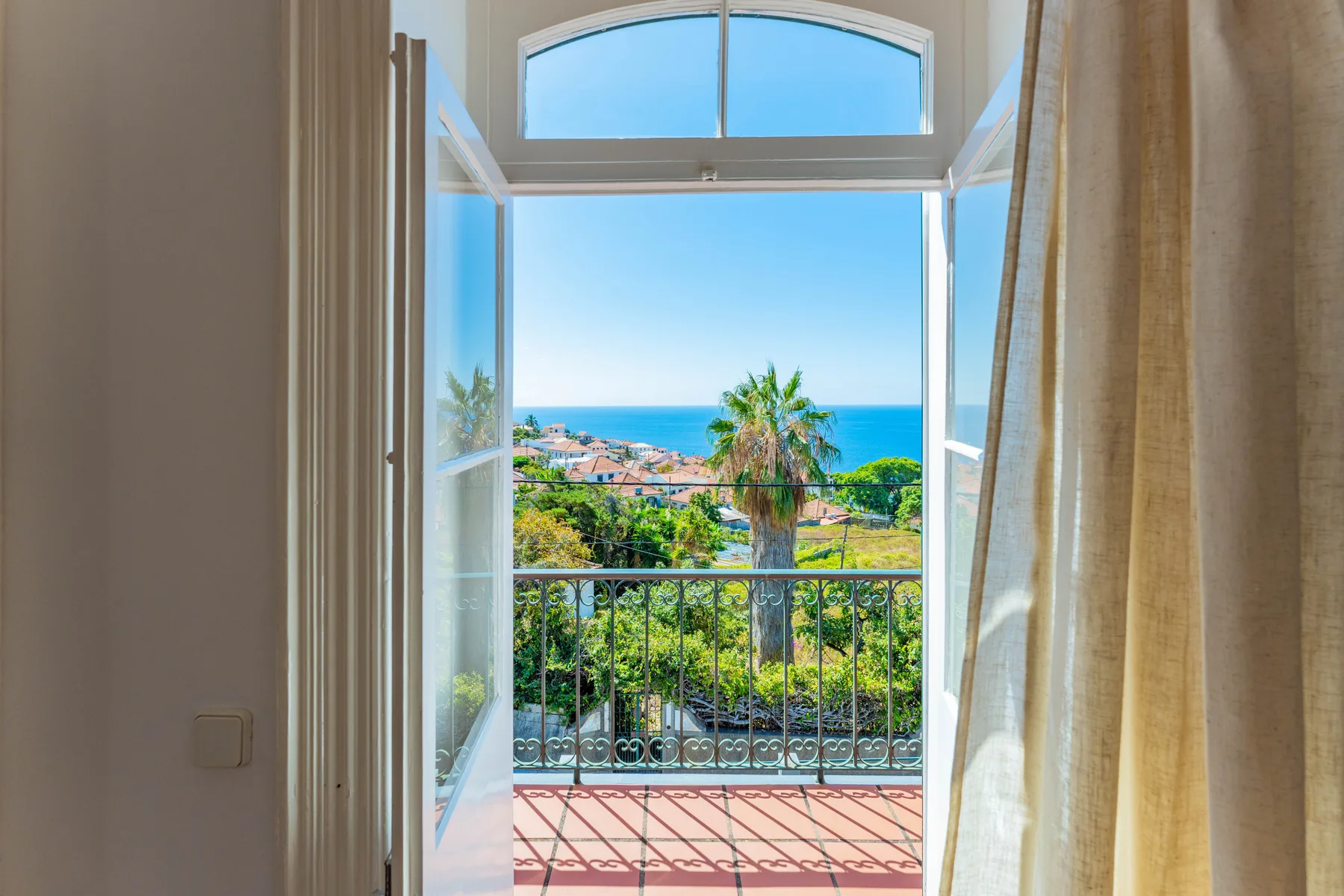 Una puerta abierta al balcón enmarca una vista de tejados, vegetación y mar.