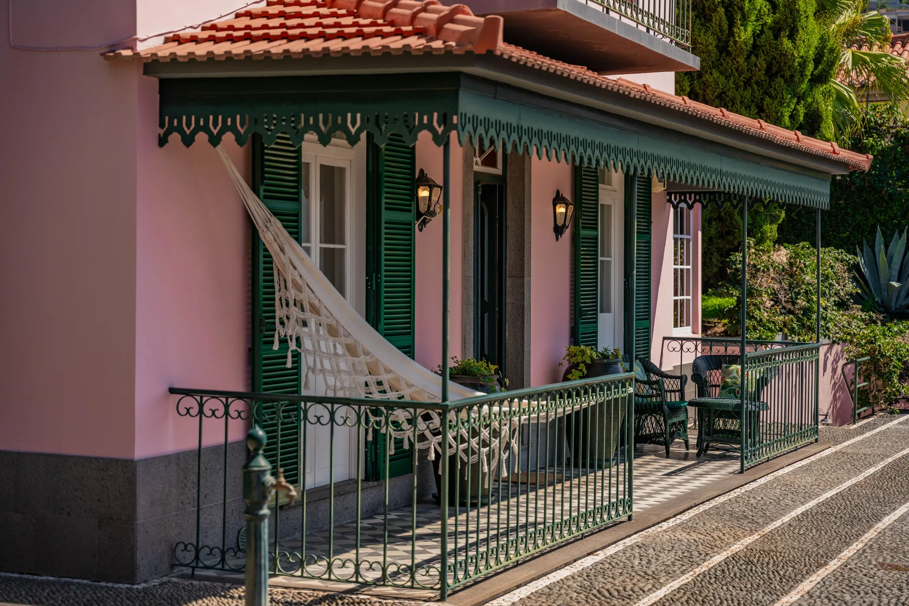 El exterior de una casa rosa tiene contraventanas verdes, barandillas ornamentadas y un sendero empedrado.