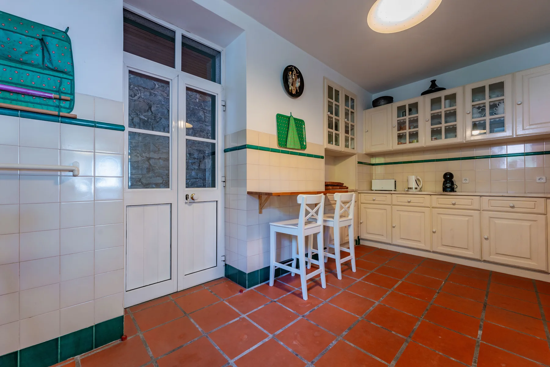 Una cocina con armarios claros, paredes alicatadas y suelo de tono terracota.
