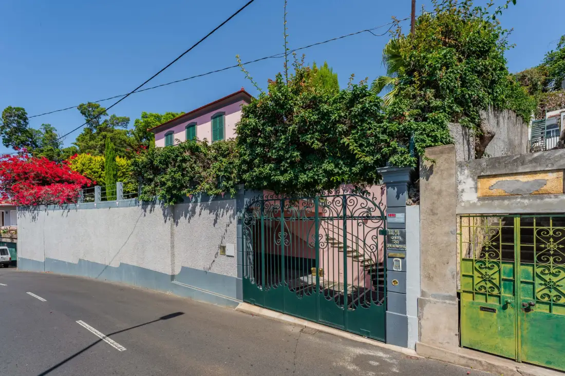 Una casa rodeada de vegetación está detrás de un muro alto y portones verdes ornamentados.