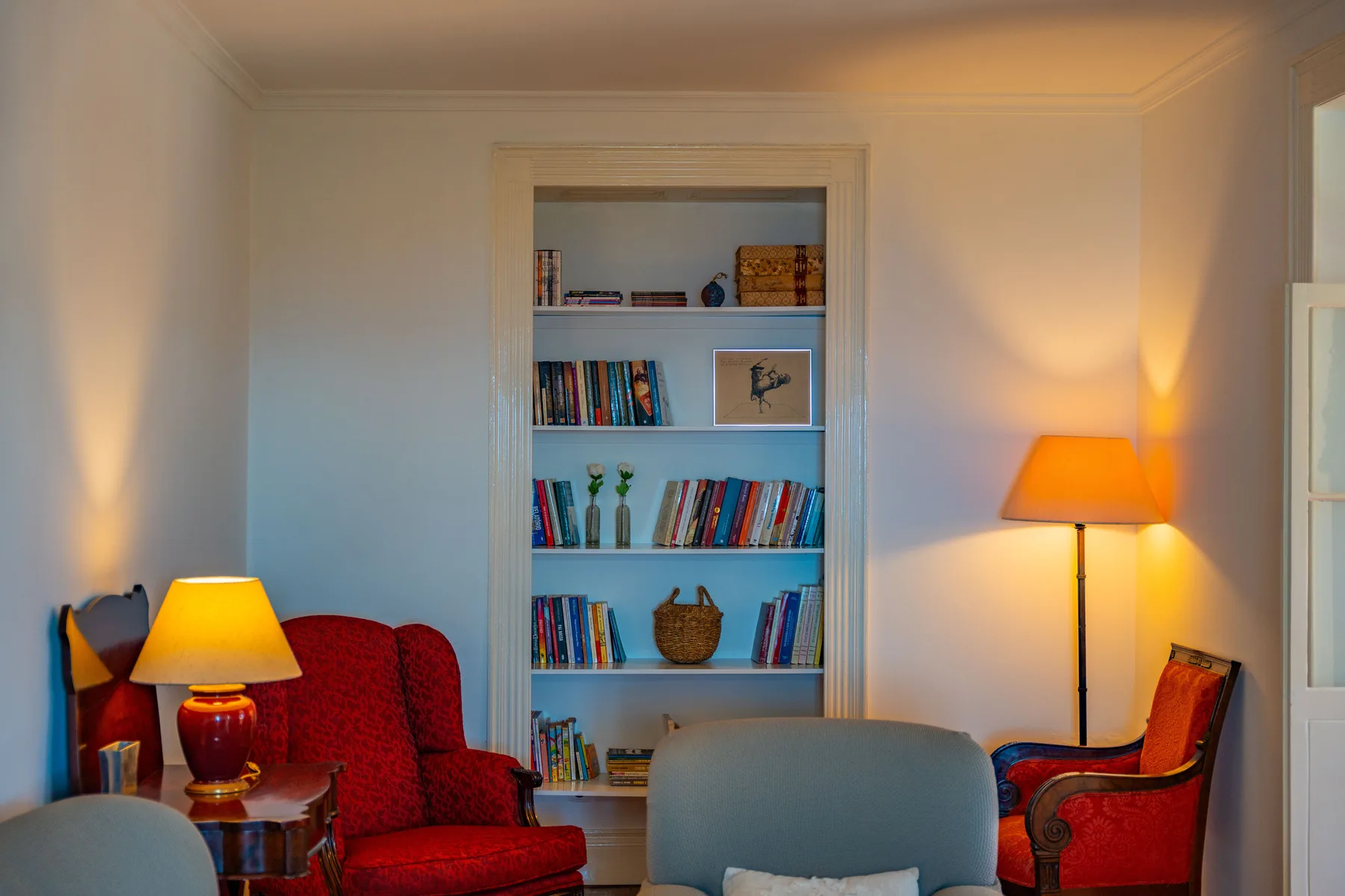 Una sala de estar con asientos rojos, una estantería y dos lámparas cálidas.