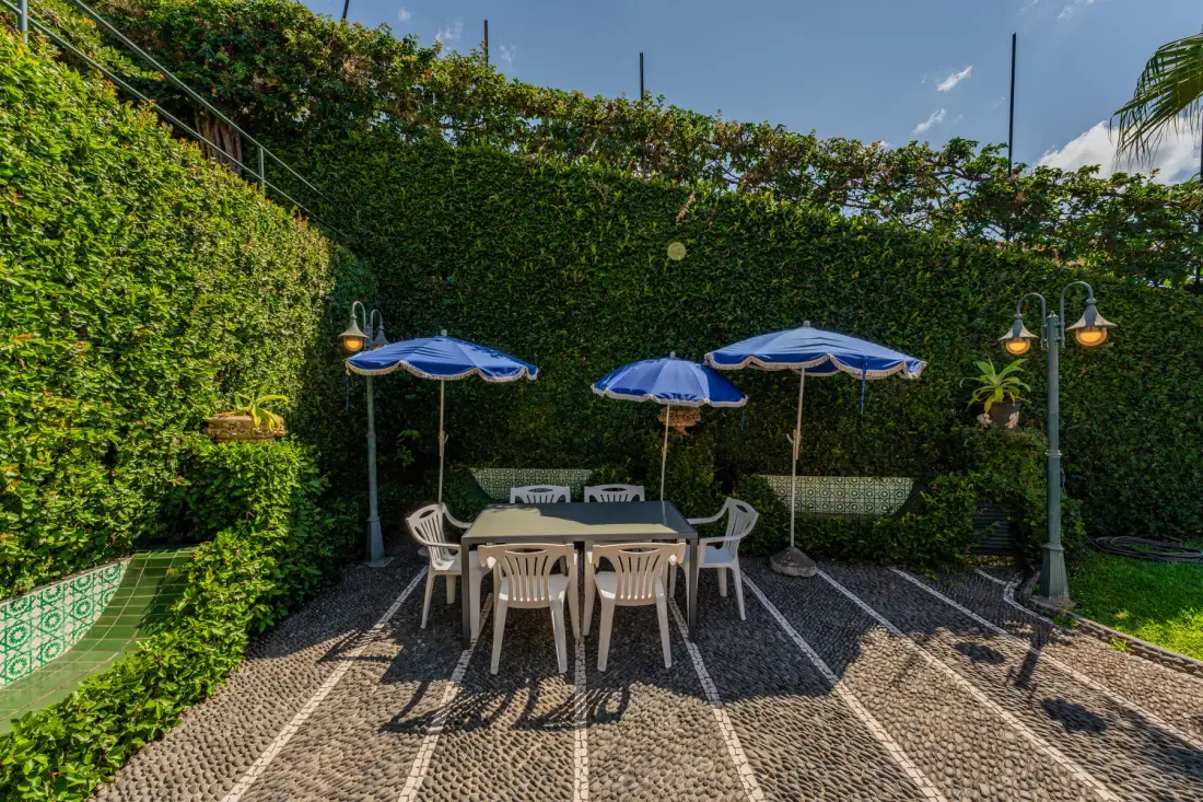 Una zona de comedor al aire libre con mesa, asientos, sombrillas y setos alrededor.