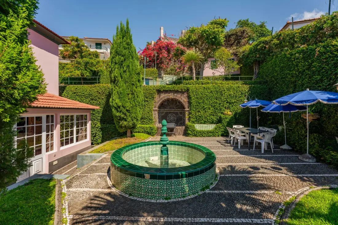 Un patio ajardinado cuenta con una fuente de azulejos, asientos al aire libre y sombrillas azules.