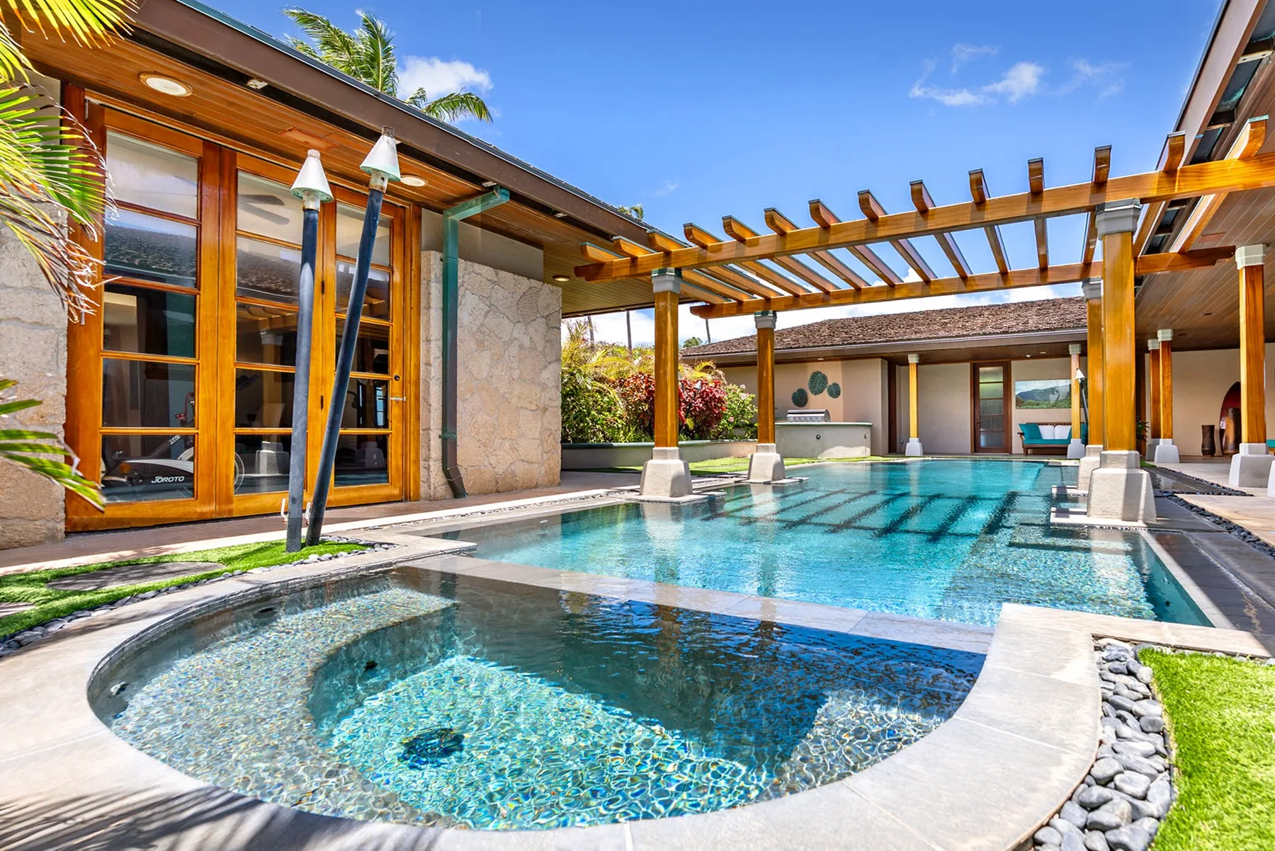 Una piscina de patio queda bajo una pérgola de madera junto a un edificio revestido de piedra.