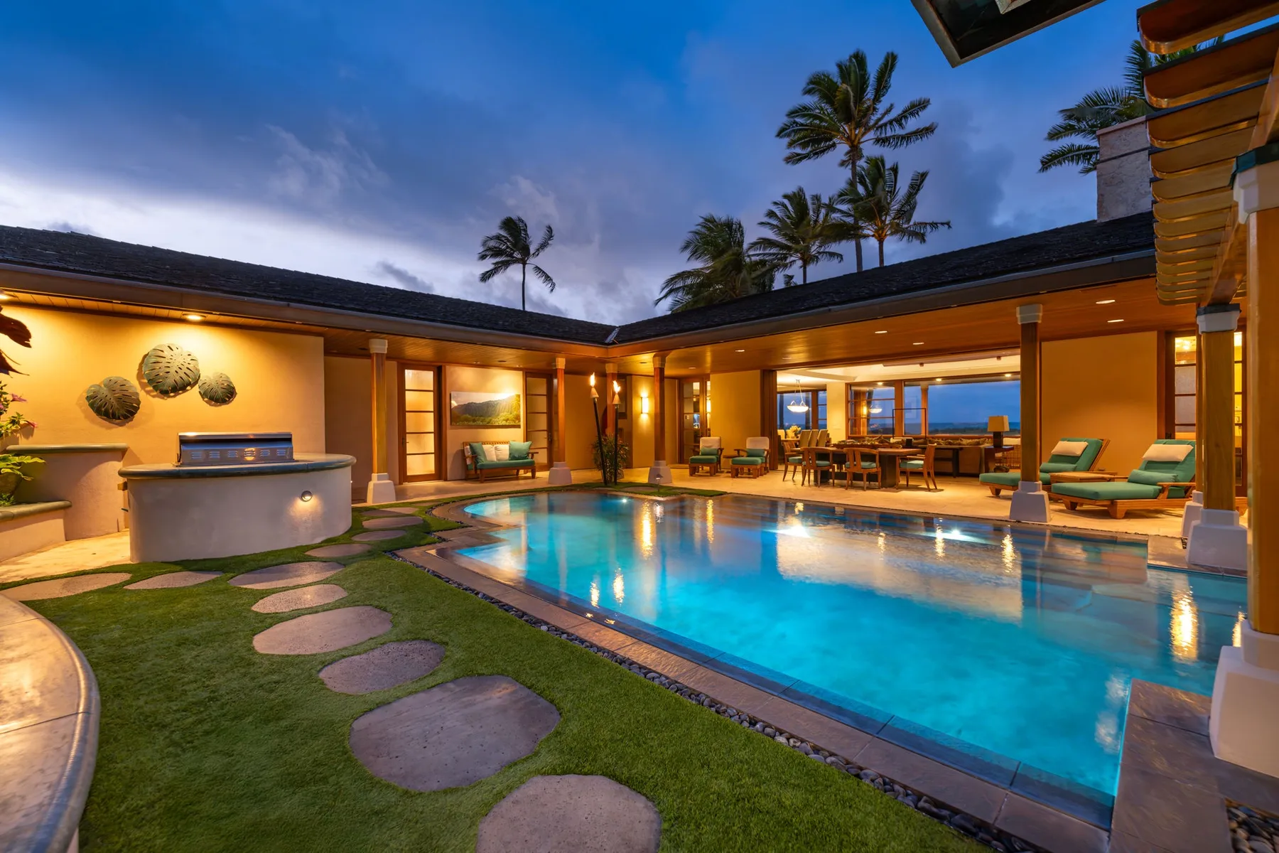 Un patio iluminado rodea una piscina, con asientos al aire libre y palmeras.