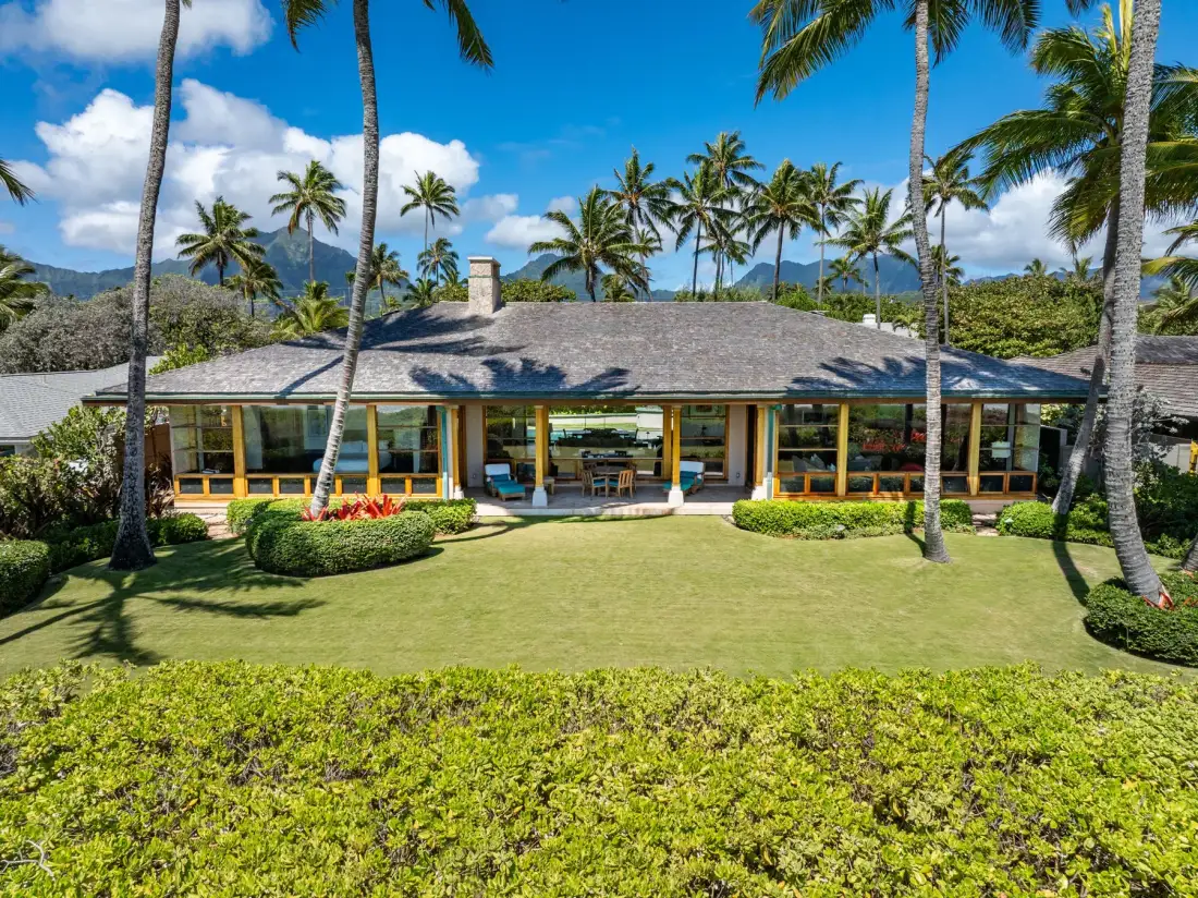 Una casa tropical con un amplio tejado se alza entre palmeras y un césped verde.