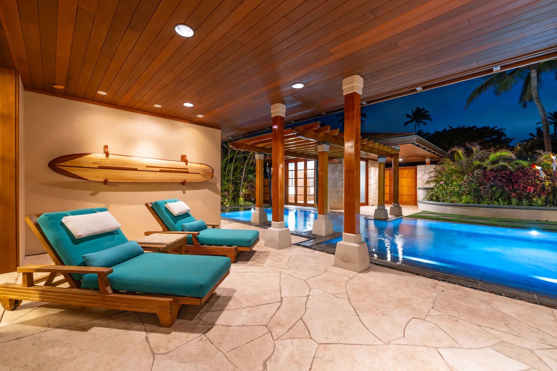 Un patio cubierto junto a la piscina tiene tumbonas turquesas junto a una piscina iluminada.