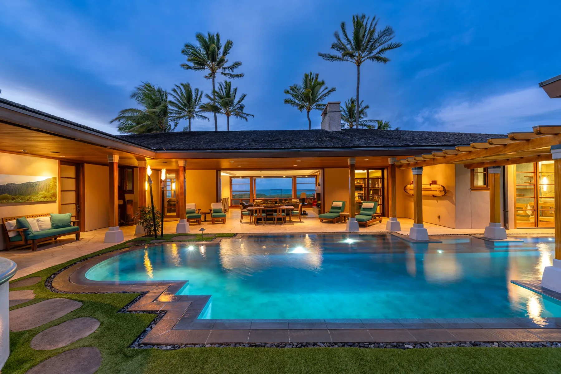 Un patio iluminado con piscina, asientos y palmeras al anochecer.