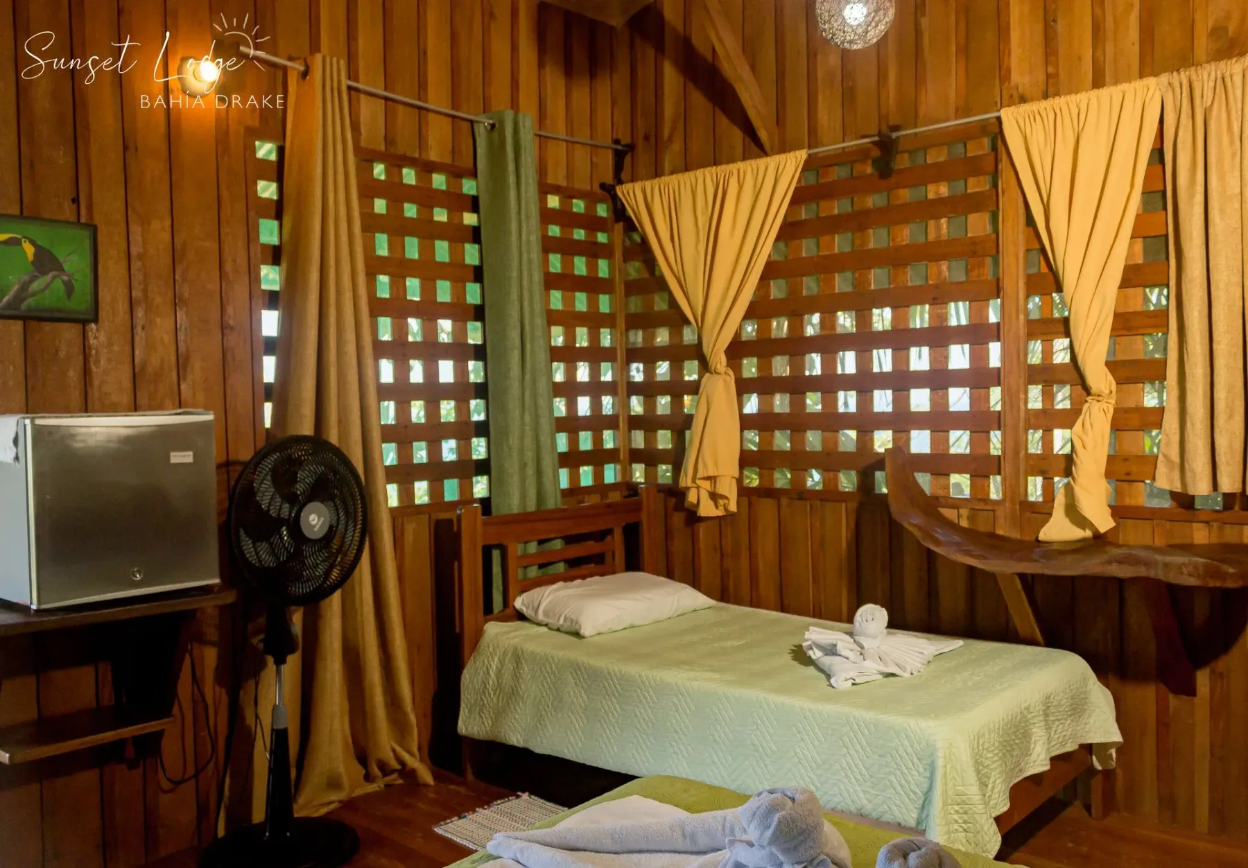 Wooden cabin bedroom with two beds, a small refrigerator, a fan, and lattice windows.