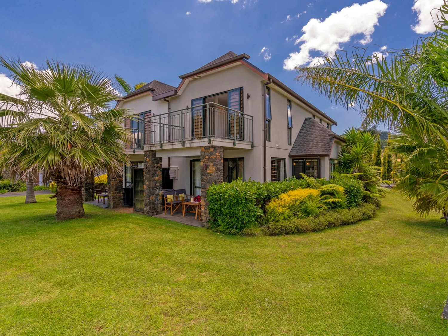 Una casa de dos plantas con balcón y patio, rodeada de palmeras y arbustos, en Villa 51 - Pauanui Holiday Home, en Pauanui.
