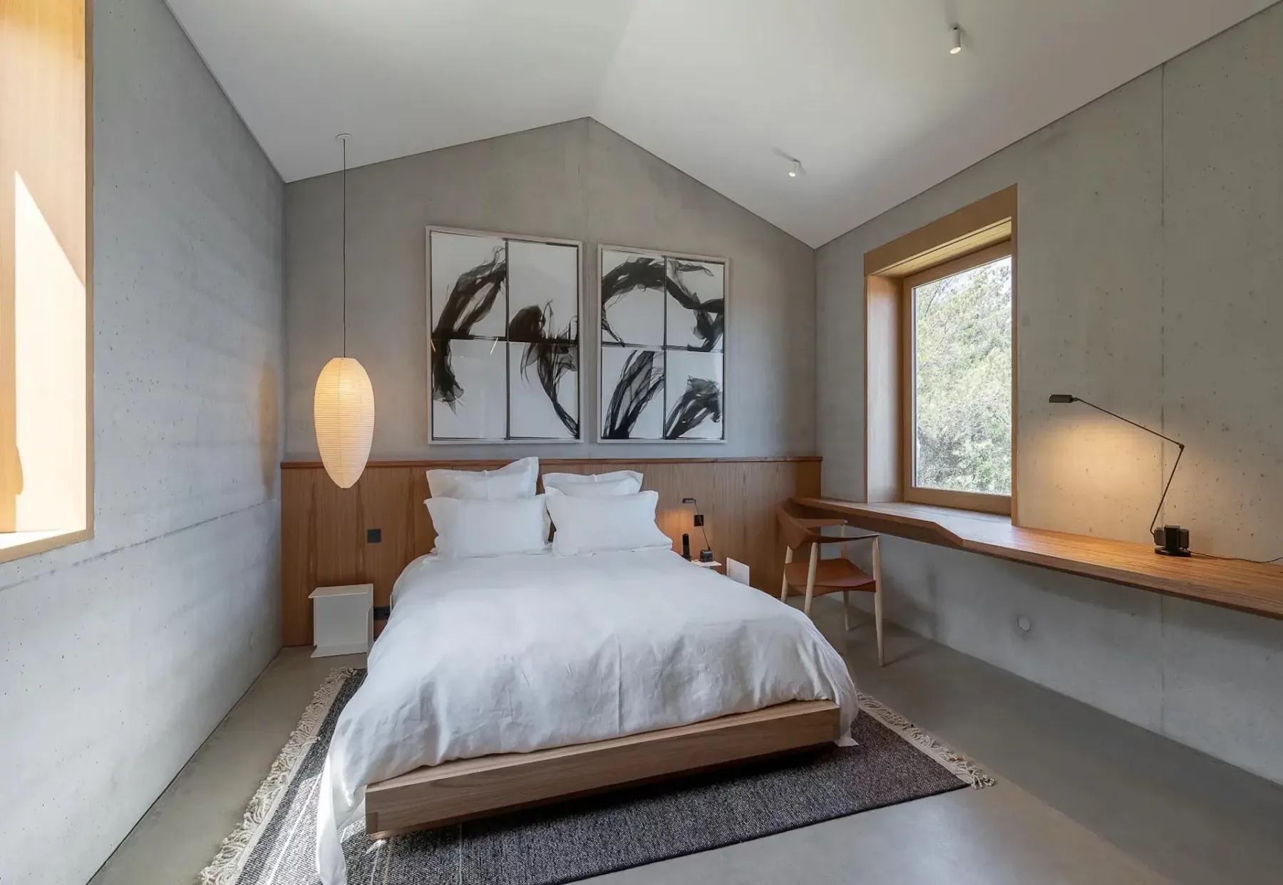 Un dormitorio moderno con una cama blanca, cabecero de madera, escritorio junto a la ventana, arte abstracto en la pared y una lámpara colgante. La habitación tiene paredes de hormigón y grandes ventanas.