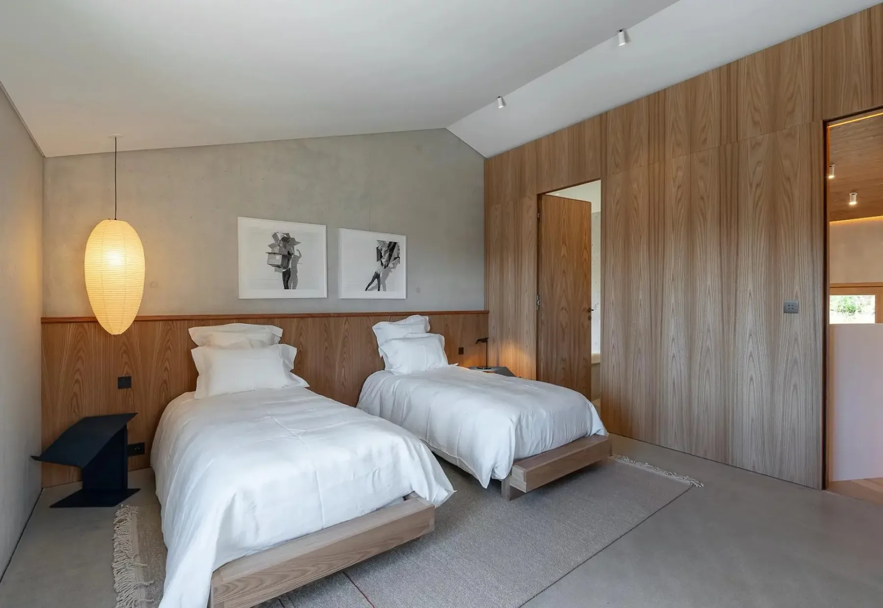 Un dormitorio moderno con dos camas individuales con ropa de cama blanca, una pared decorativa de madera, una lámpara colgante grande, obras de arte minimalistas en las paredes y una alfombra en el suelo.