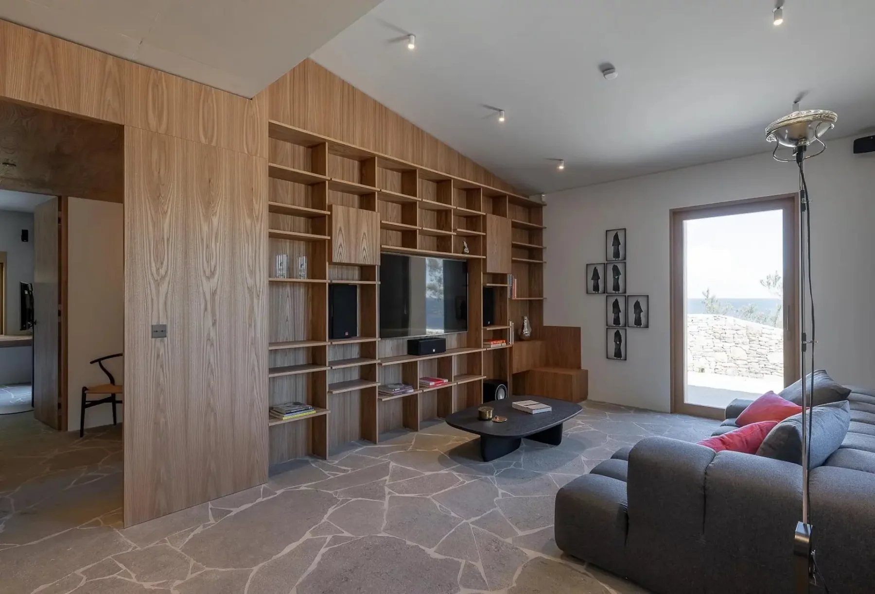 Una sala de estar moderna con un mueble de pared de madera, estantes integrados, un televisor, un sofá, una mesa baja y fotografías enmarcadas en la pared. Una puerta de cristal da al exterior. Se aprecia el suelo de piedra.