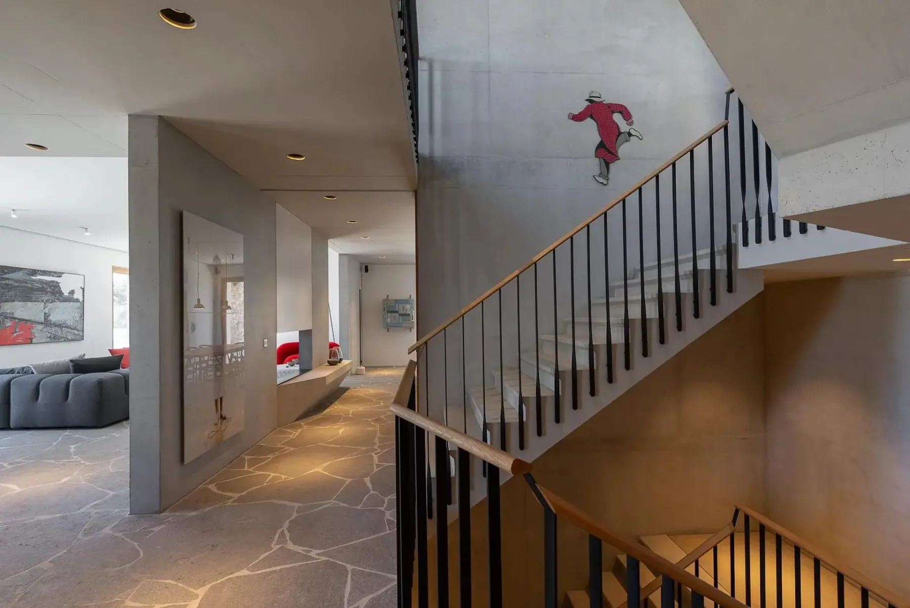 Un interior moderno con suelo de piedra, paredes de hormigón y una escalera con barandillas negras. Sobre la escalera se ve una obra de arte mural en rojo y blanco. Al fondo hay un sofá gris y piezas de arte.