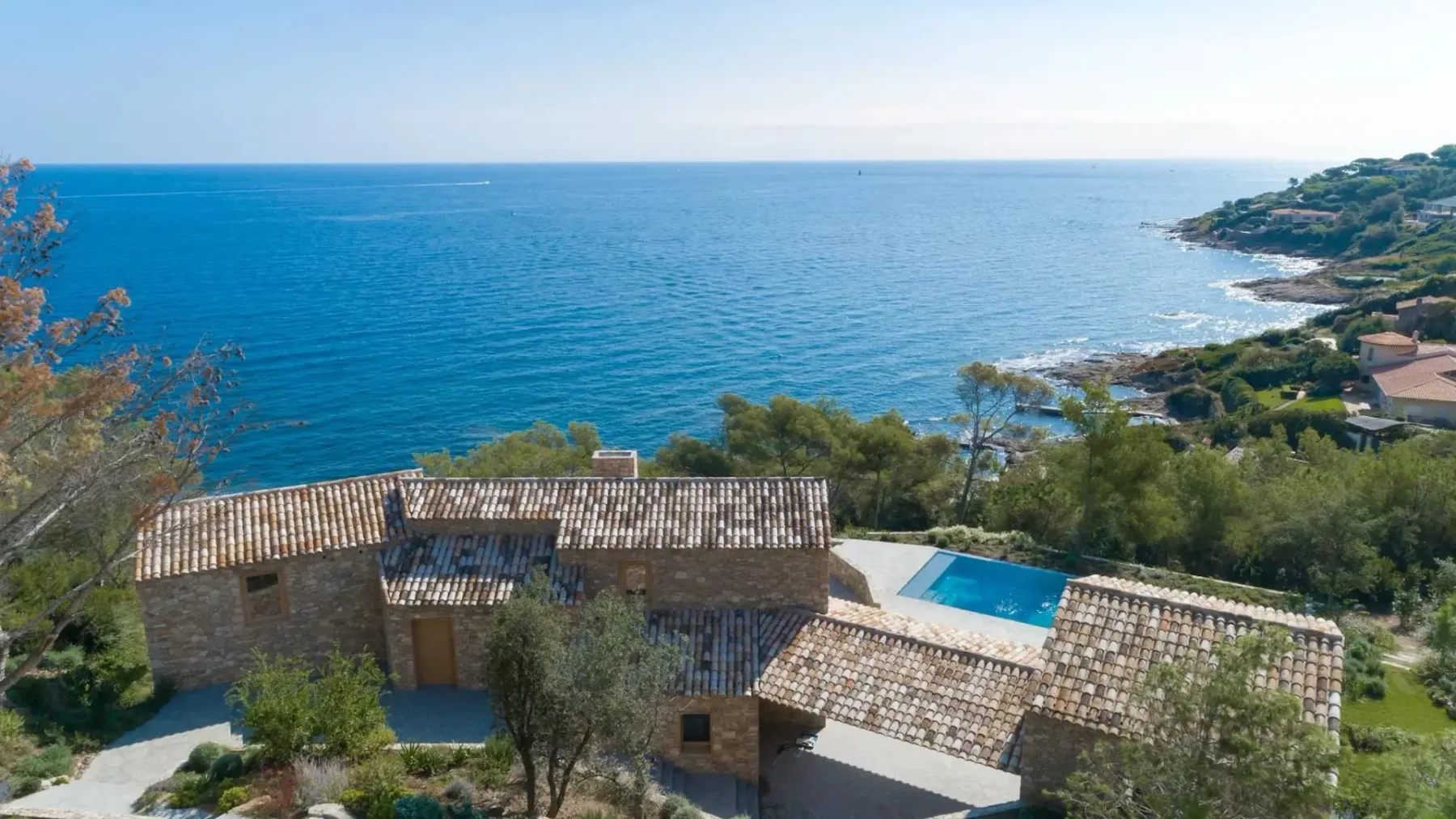Una casa de piedra con tejado de tejas y piscina tiene vistas a una costa de aguas azules y formaciones terrestres lejanas en un día despejado.