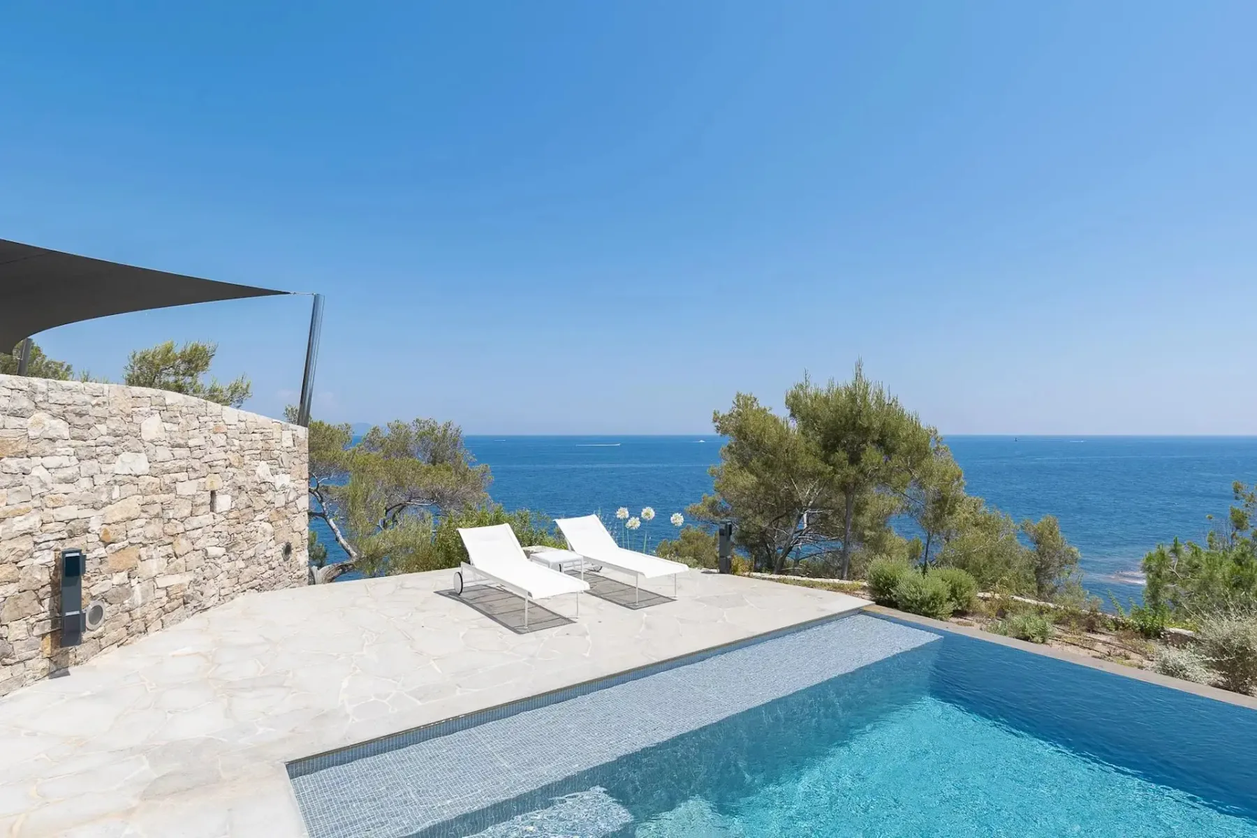 Una piscina infinita junto al mar con dos tumbonas blancas, con vistas a un océano azul de aguas cristalinas, rodeada de vegetación y con un muro de piedra a un lado.