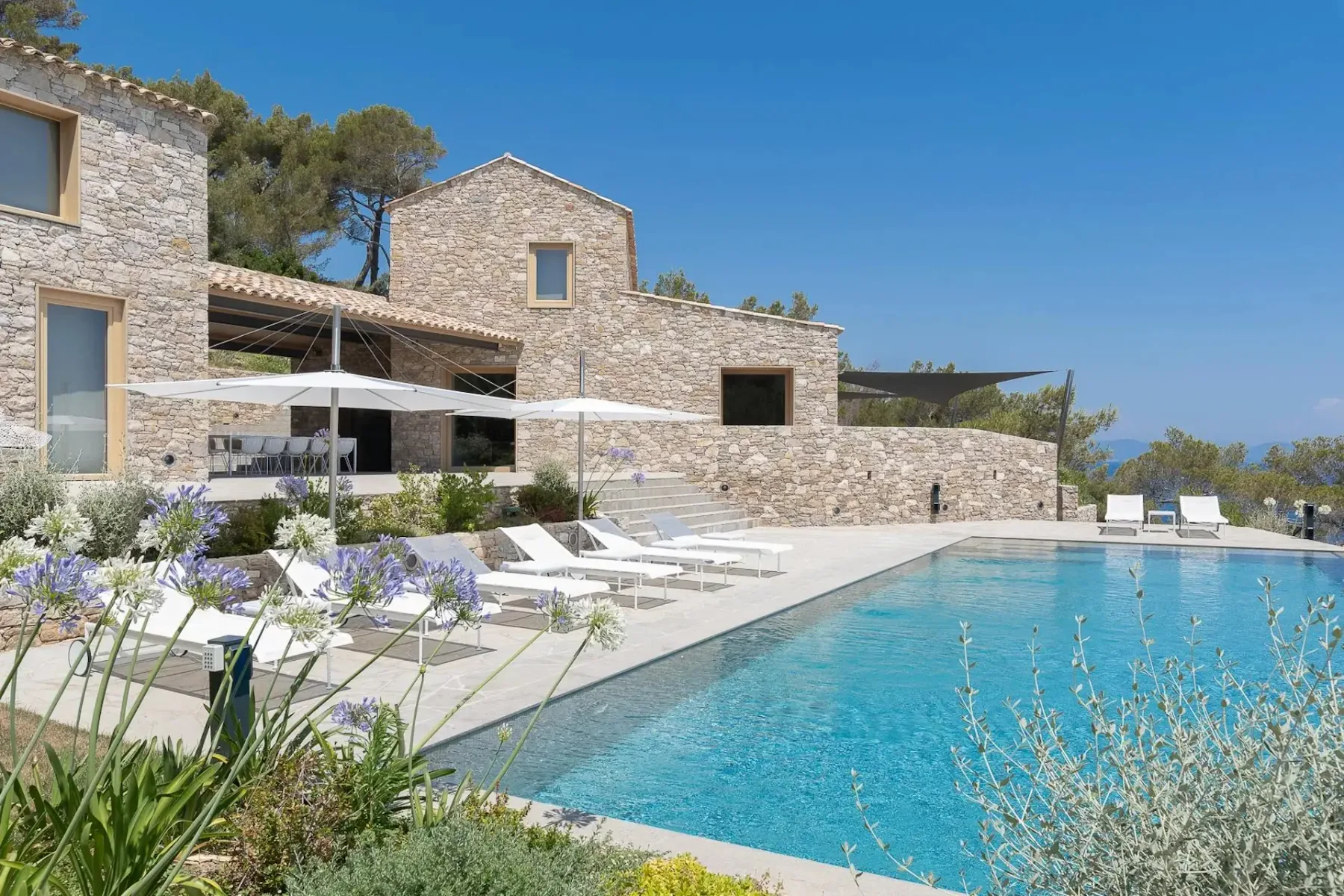 Una casa de piedra de varios niveles tiene una gran piscina delante, rodeada de tumbonas y sombrillas. La zona está ajardinada con diversas plantas y el día es soleado, con el cielo despejado.