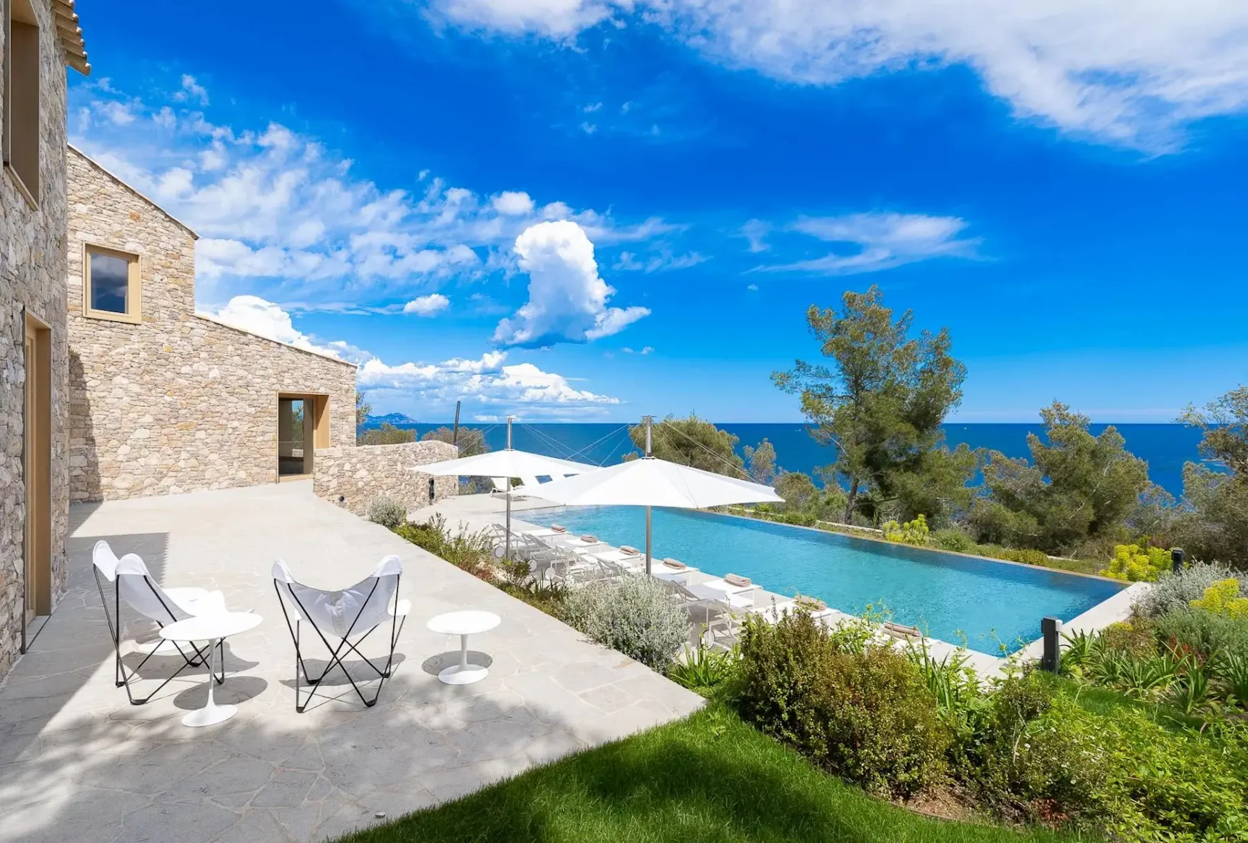 Un patio de piedra con muebles blancos tiene vistas a una piscina infinita y al océano, rodeado de vegetación y bajo un cielo azul luminoso con nubes.