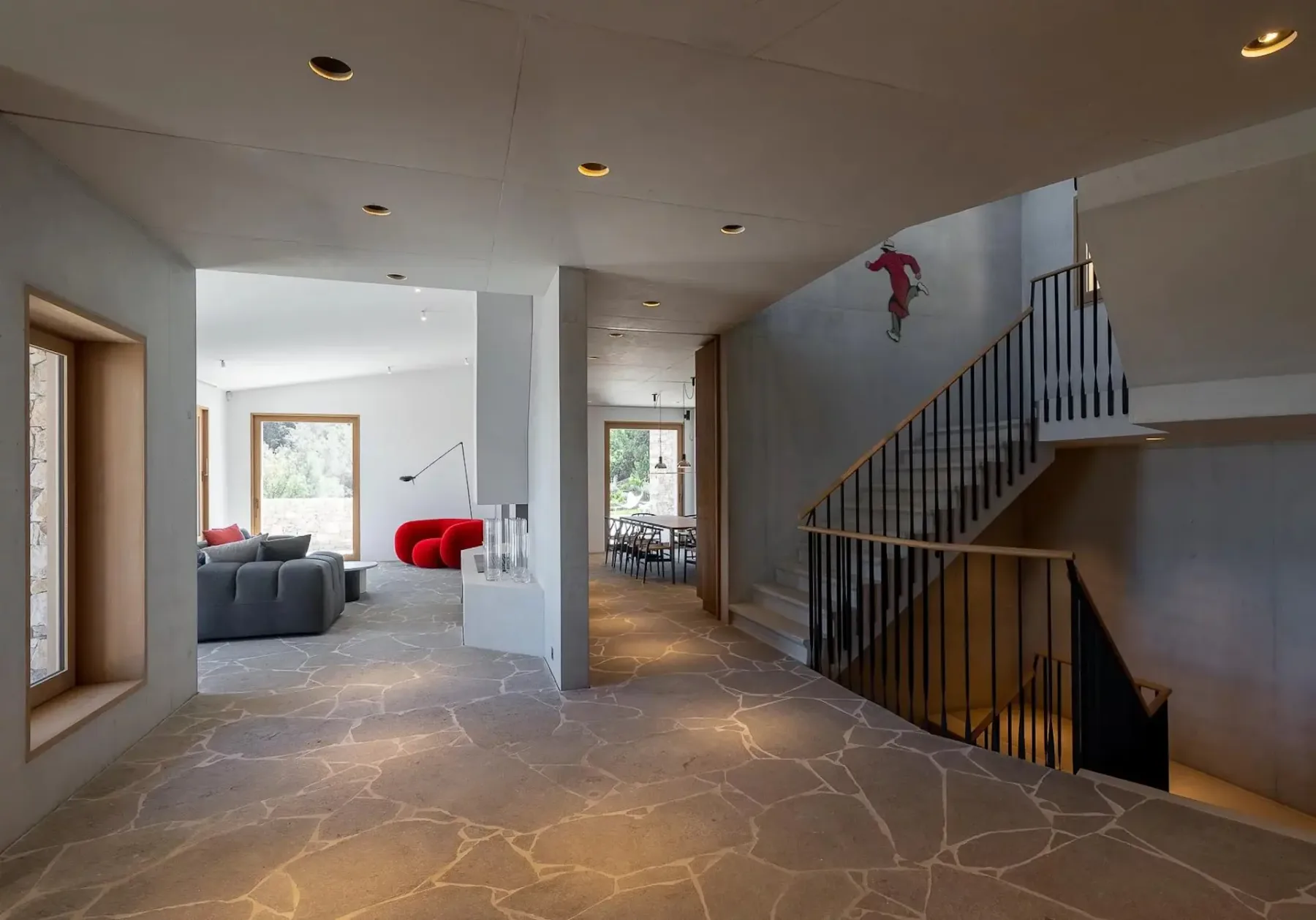 Un interior moderno con un pasillo con suelo de piedra, una escalera, una zona de estar con muebles grises y rojos y grandes ventanas que dejan entrar luz natural.