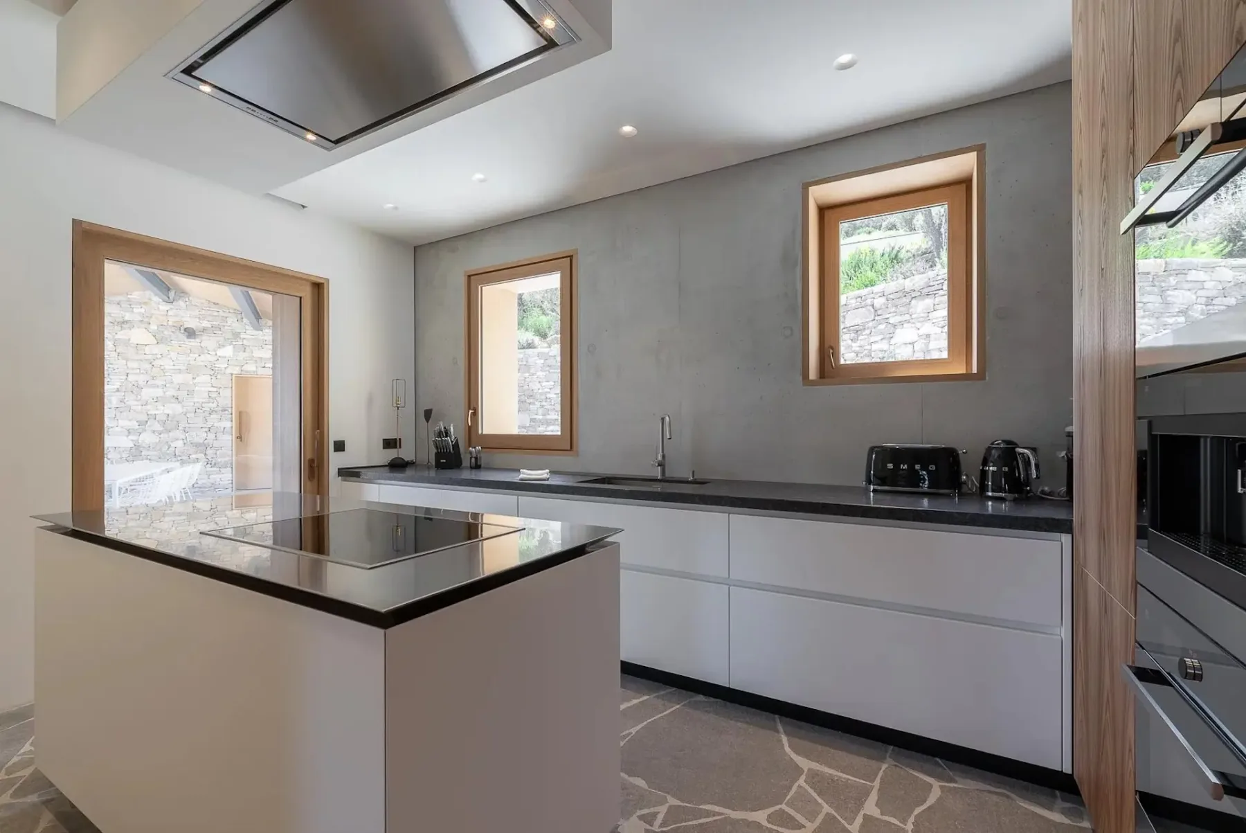 Una cocina moderna con una isla central, placa de inducción y campana extractora de acero inoxidable, que cuenta con dos ventanas, una pared de hormigón y electrodomésticos contemporáneos.
