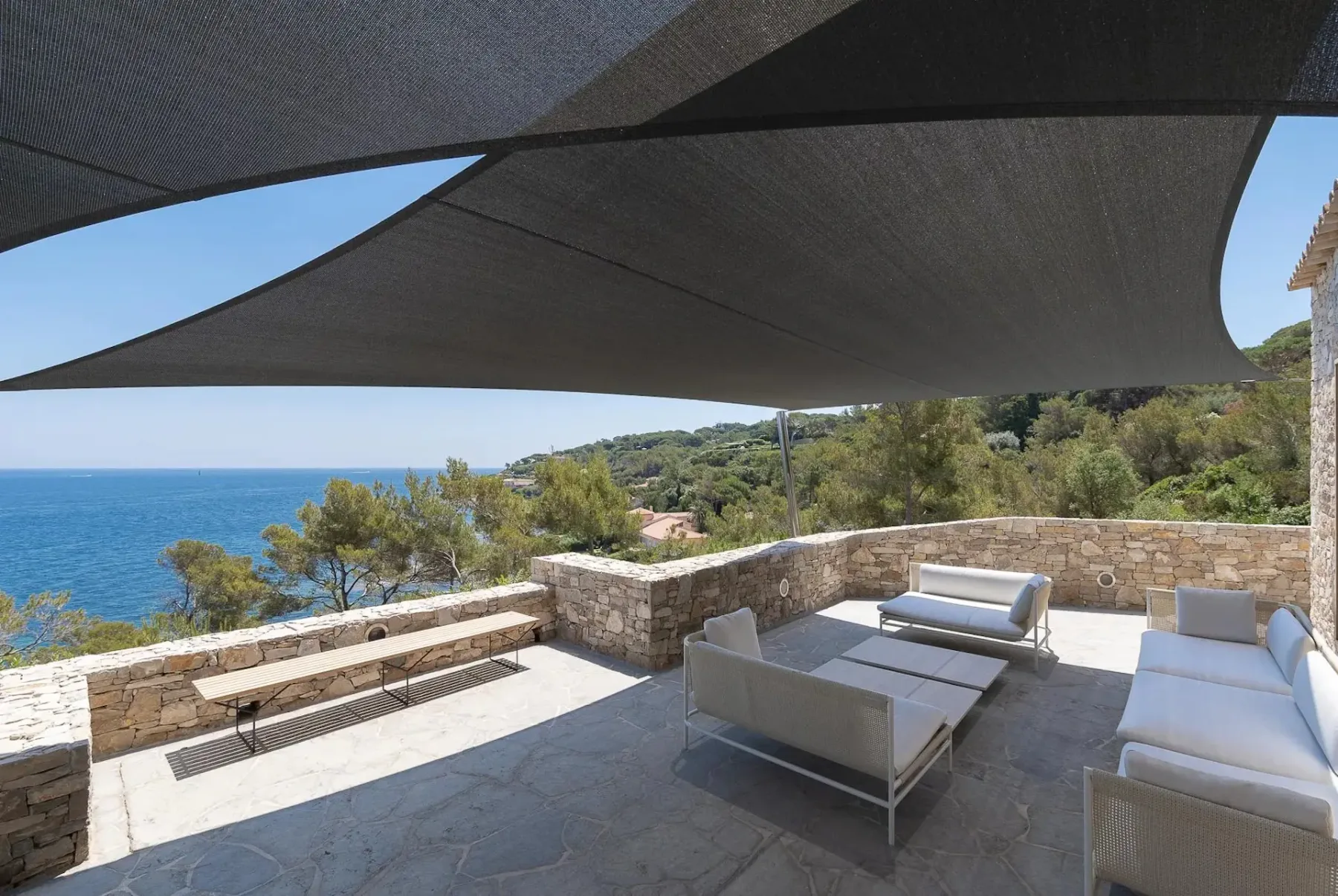 Patio exterior con paredes de piedra, muebles blancos modernos, un banco y toldos superiores, con vistas al océano azul y a una ladera verde.