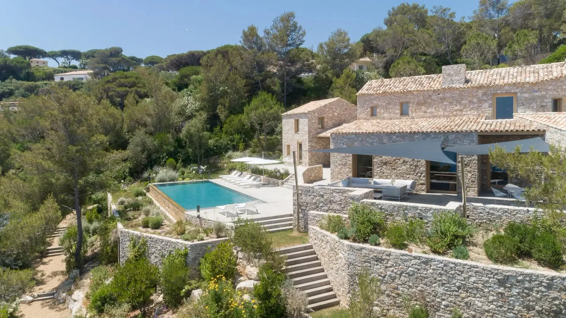 Una casa de paredes de piedra con varias secciones y una piscina al aire libre, rodeada de árboles y vegetación, situada en una ladera con un patio y zonas de estar exteriores.