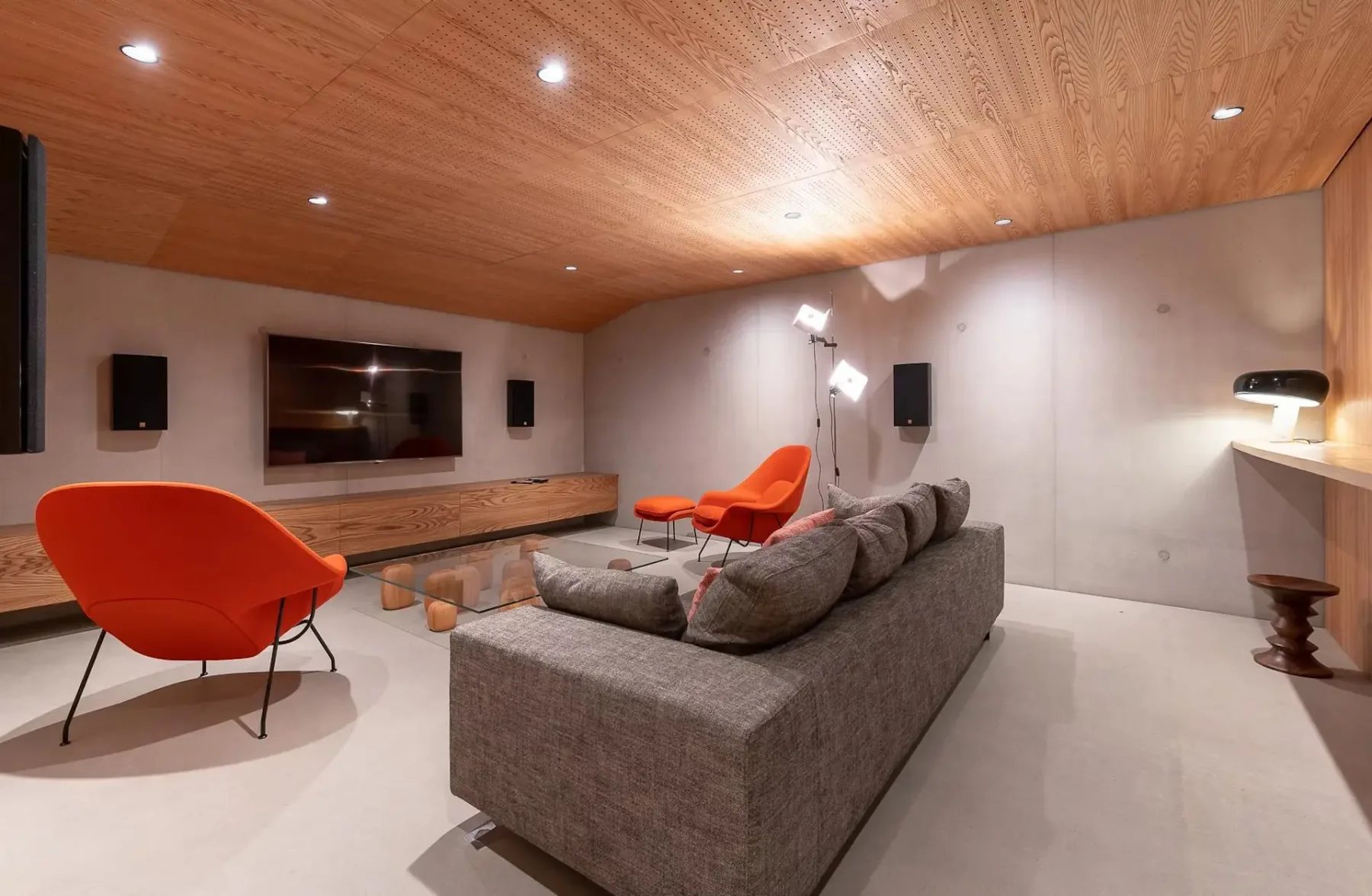 Una sala de estar moderna con asientos grises, sillas naranjas y un televisor montado en la pared.