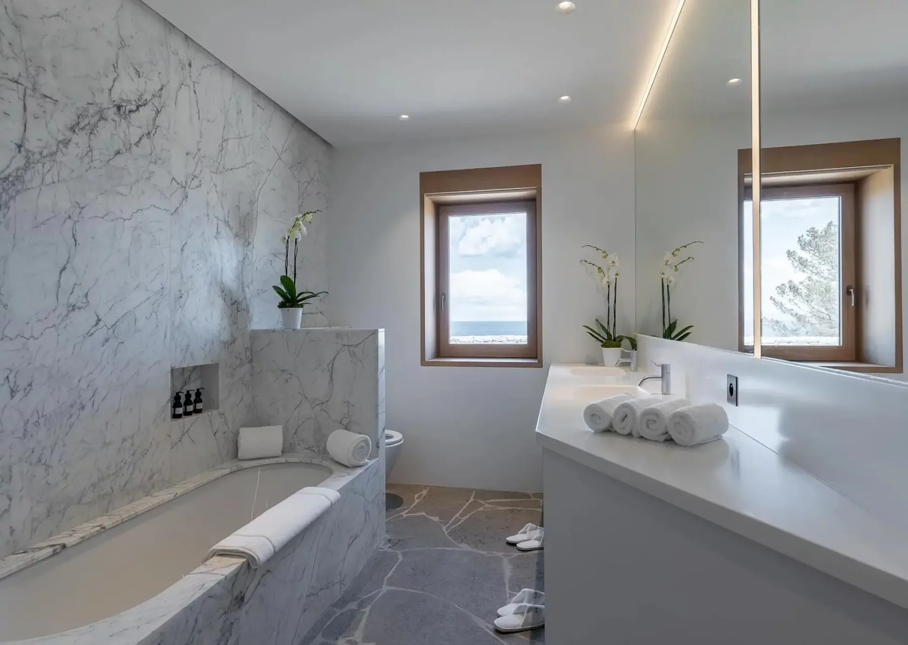 Un baño moderno con paredes y suelo de mármol cuenta con una bañera, un espejo grande, un lavabo con toallas, dos orquídeas y una ventana con vistas al exterior.