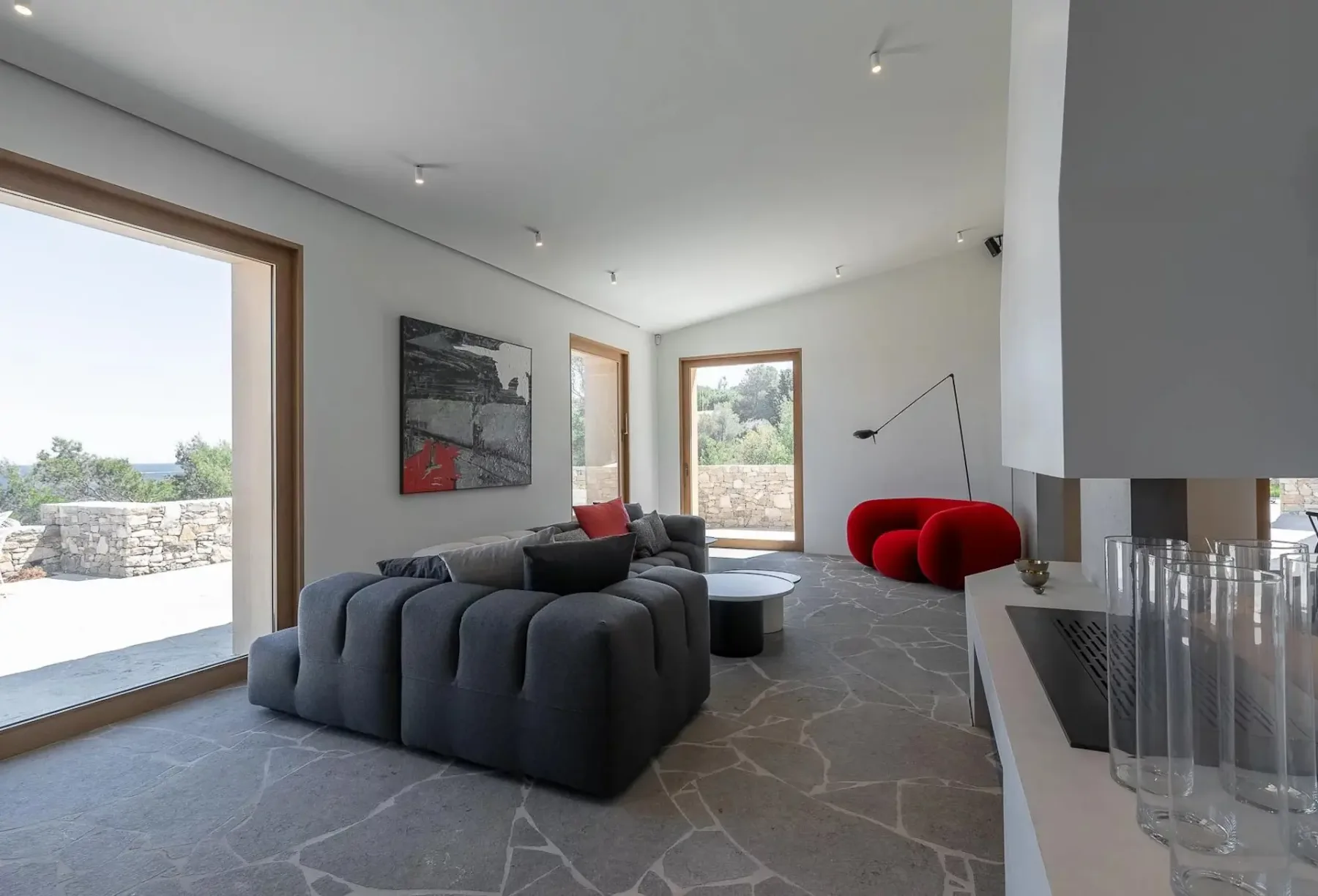 Una sala de estar moderna con grandes ventanas, un sofá modular gris, sillas rojas, un televisor montado en la pared y arte en la pared. La estancia tiene suelo con textura de piedra y vistas a un muro de piedra y a la vegetación exterior.