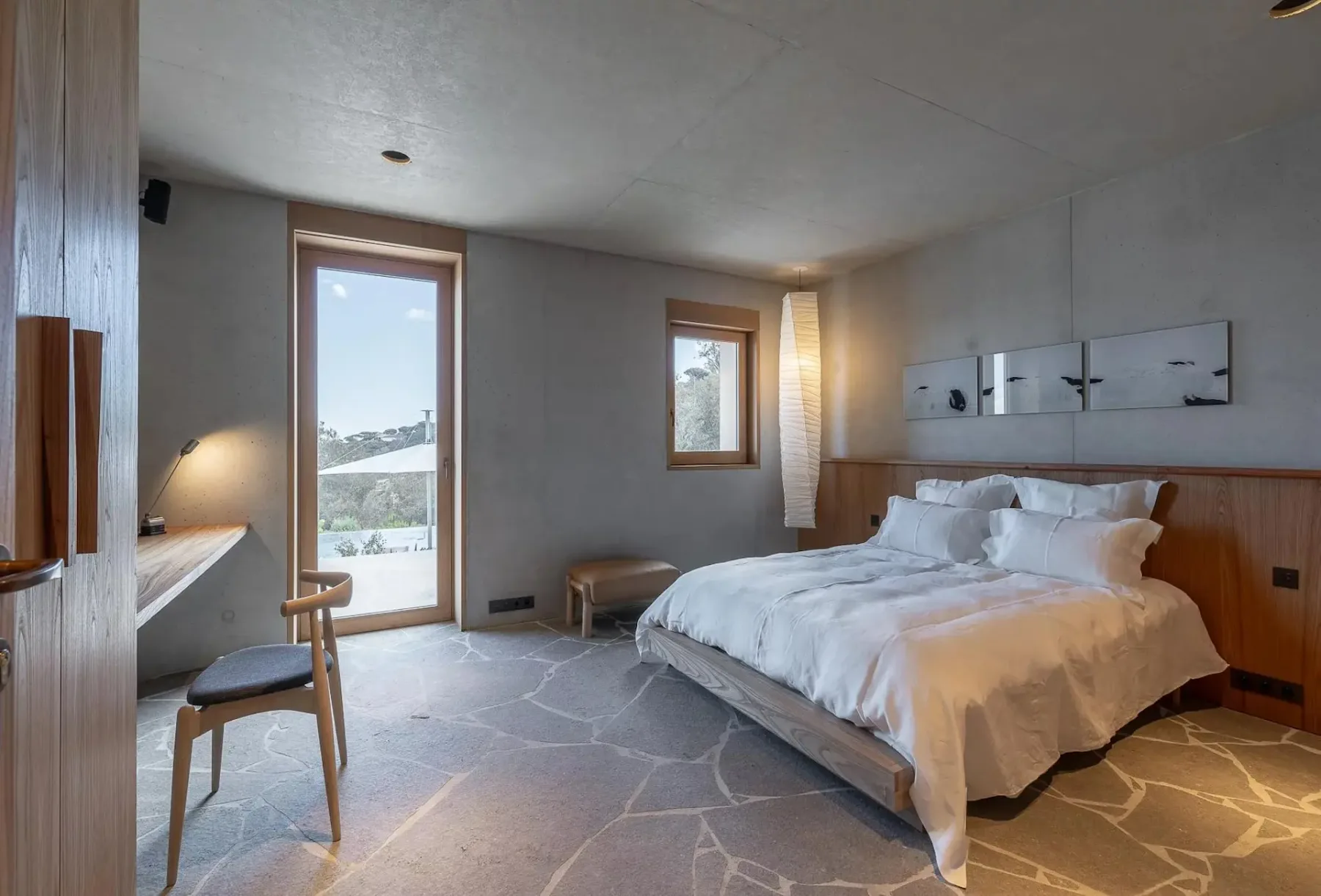 Dormitorio moderno con una ventana grande, cama doble con ropa de cama blanca, detalles de madera, un escritorio, una silla y decoración minimalista. La luz natural ilumina la habitación.