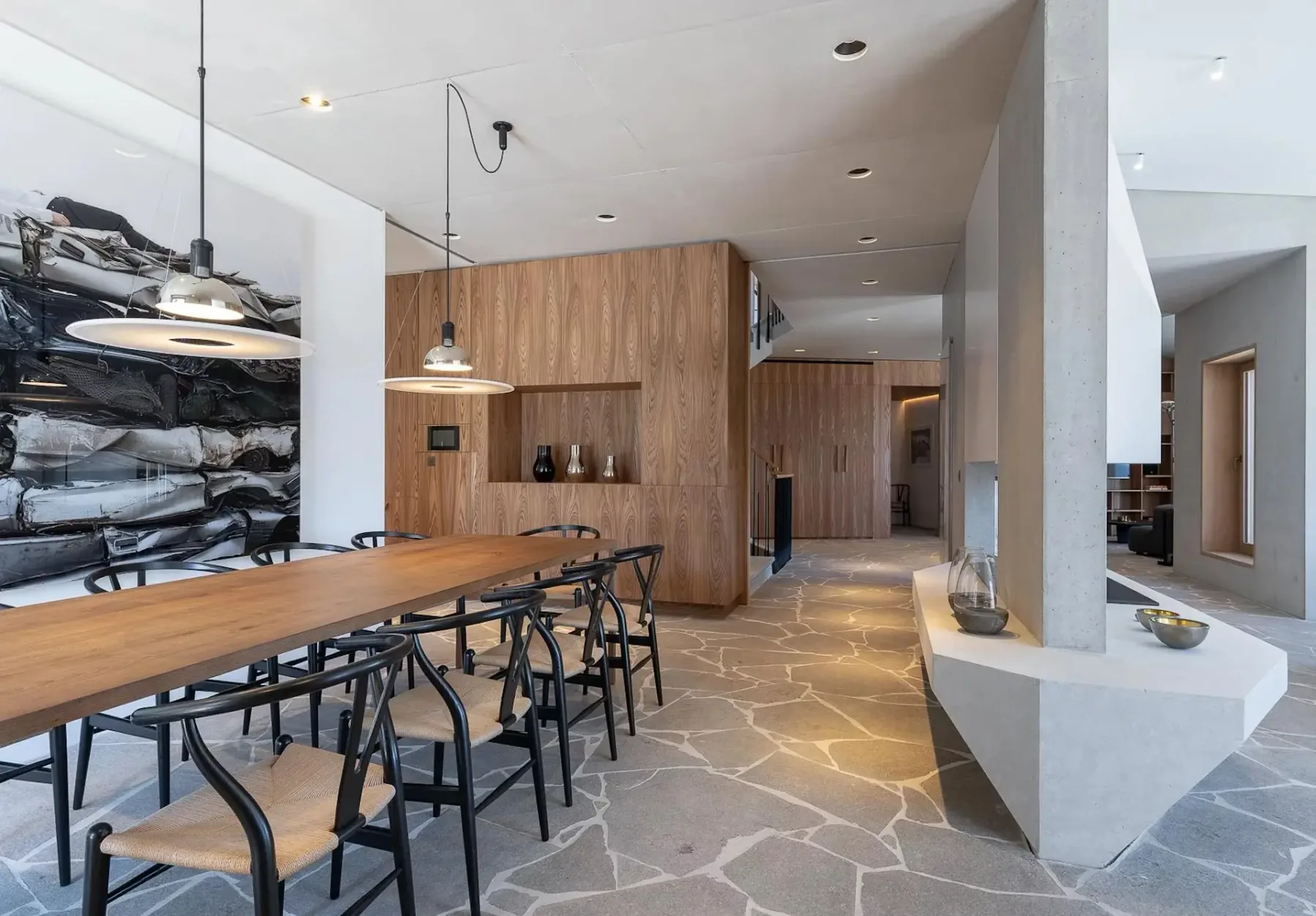 Comedor moderno con una mesa larga de madera, sillas negras, lámparas colgantes y suelo de piedra. El espacio cuenta con revestimiento de madera, una estantería integrada y elementos decorativos minimalistas.