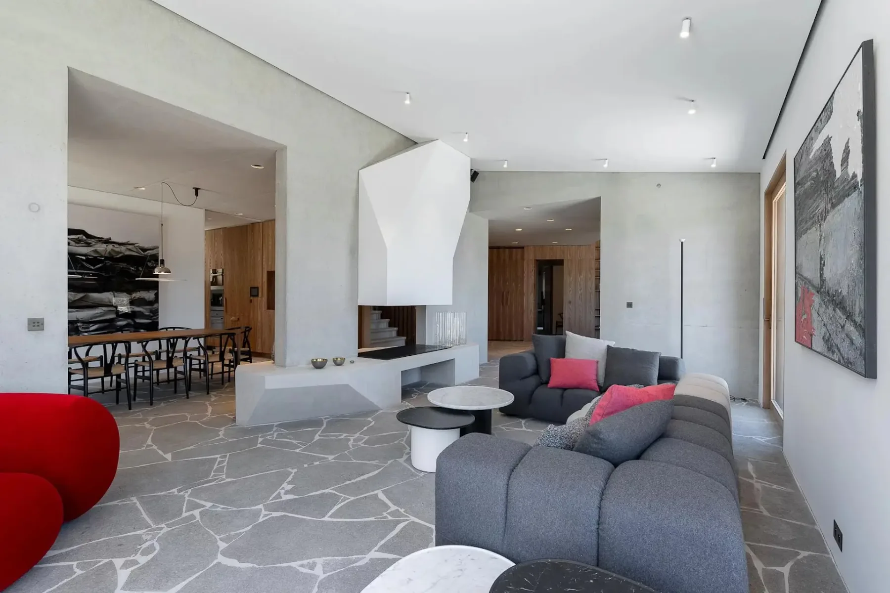 Sala de estar moderna con sofá modular gris, suelo de piedra con diseño geométrico, sillas decorativas rojas, arte abstracto en la pared y vistas al comedor contiguo.