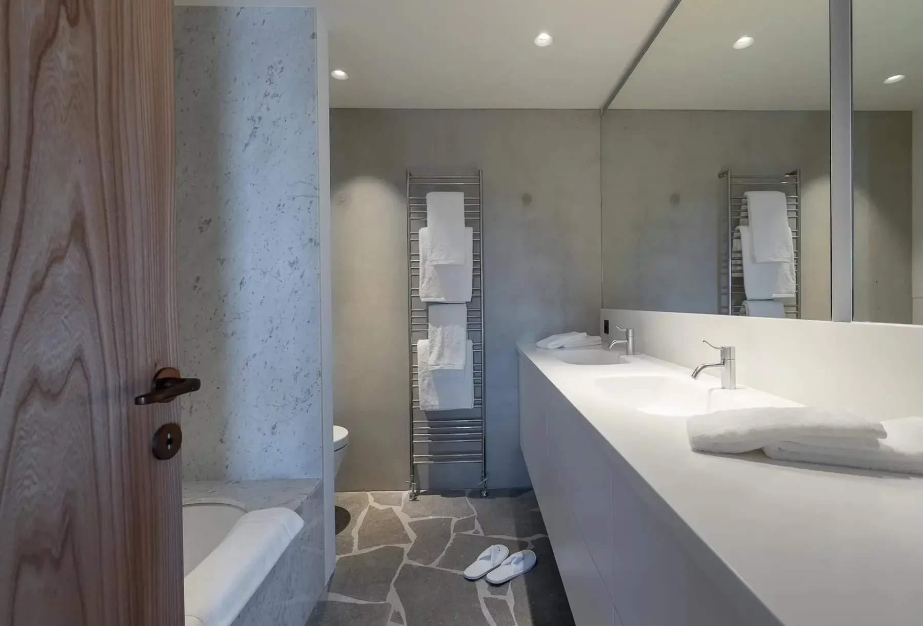 Baño moderno con encimera de doble lavabo, espejo grande, luces superiores encendidas, zona de ducha con cristal esmerilado, toallero y toallas blancas. El suelo tiene un diseño de baldosas de piedra y hay zapatillas cerca.