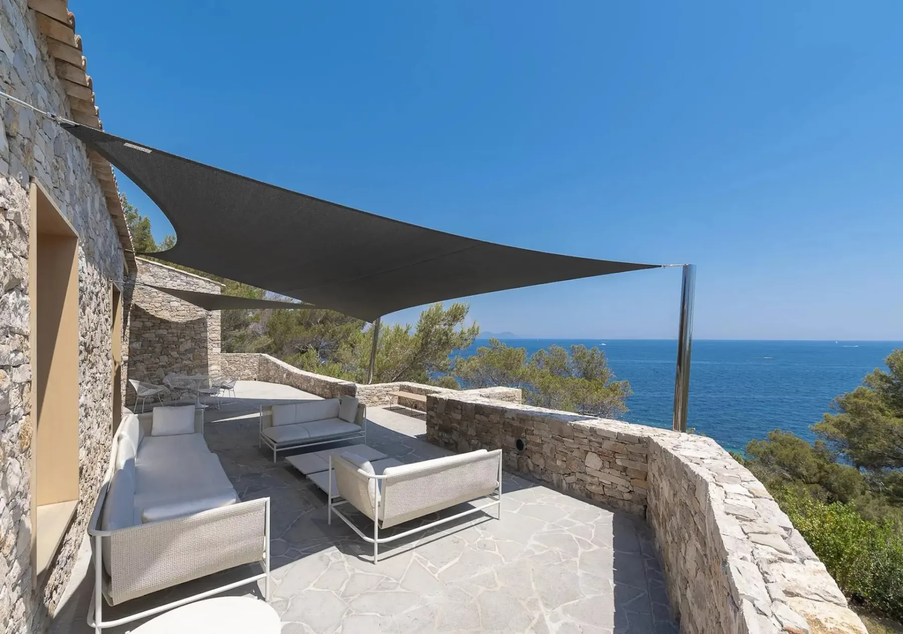 Patio exterior con asientos blancos y un toldo de vela, con vistas al mar en un día soleado de cielo azul despejado. El patio está rodeado de paredes de piedra.